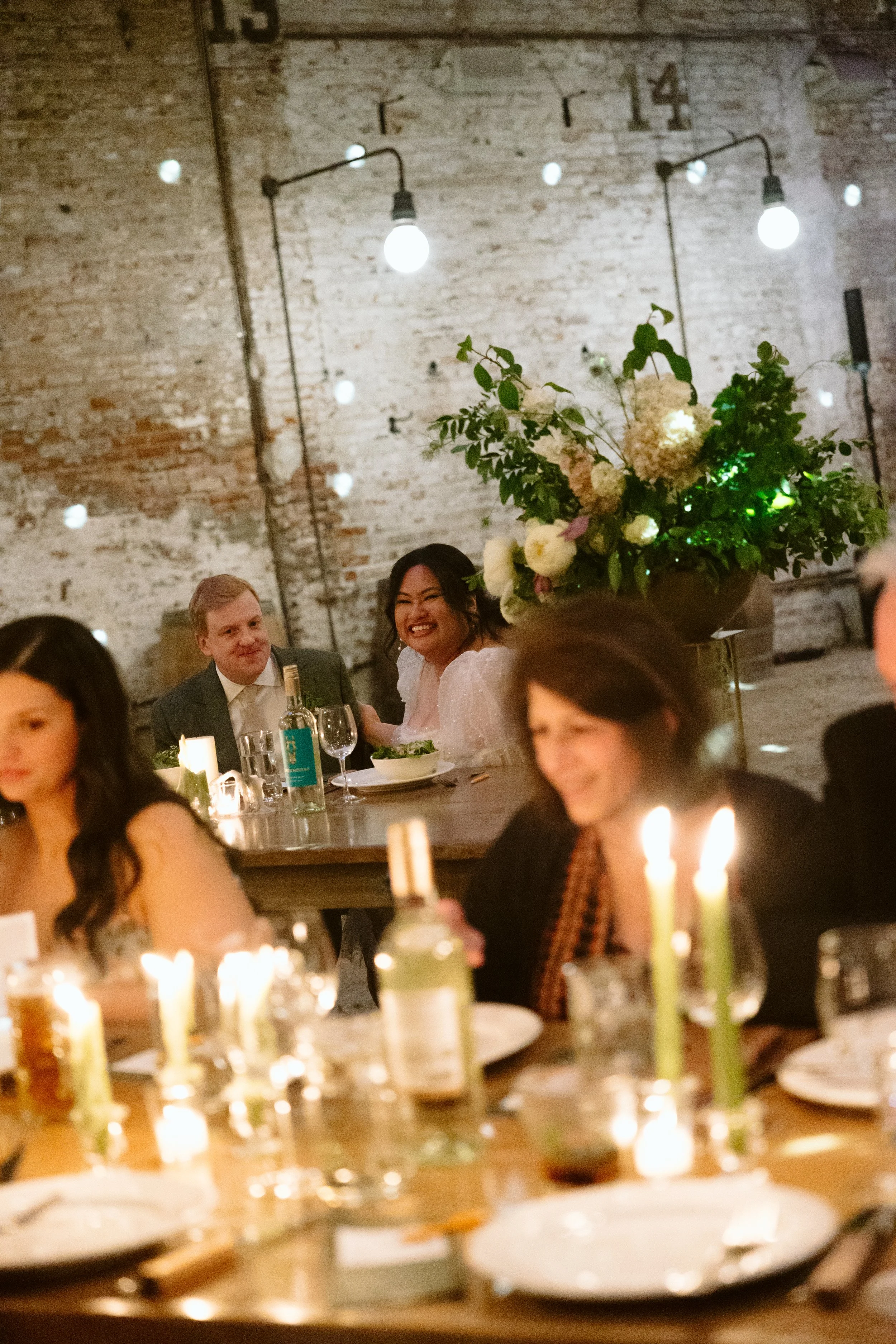 People at a dinner party with candles, a large floral arrangement, and exposed brick walls, celebrating in a rustic venue. marigolds florals nyc florist wedding florals houston hall nyc bar wedding