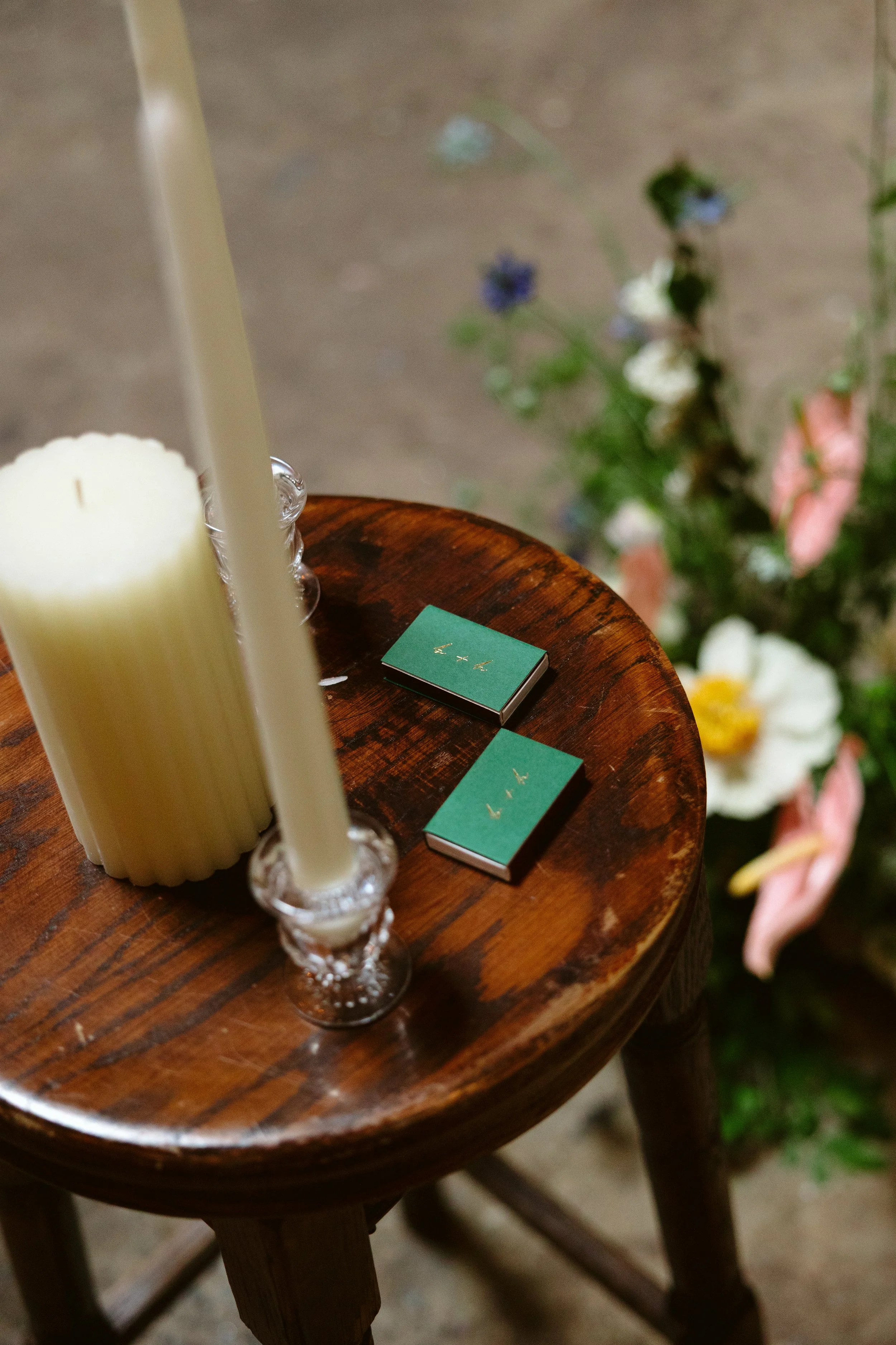 Close-up of a round wooden table with two candles in glass holders and two small teal boxes with gold writing on top, with a blurred background of flowers. marigolds florals nyc florist wedding florals houston hall nyc bar wedding