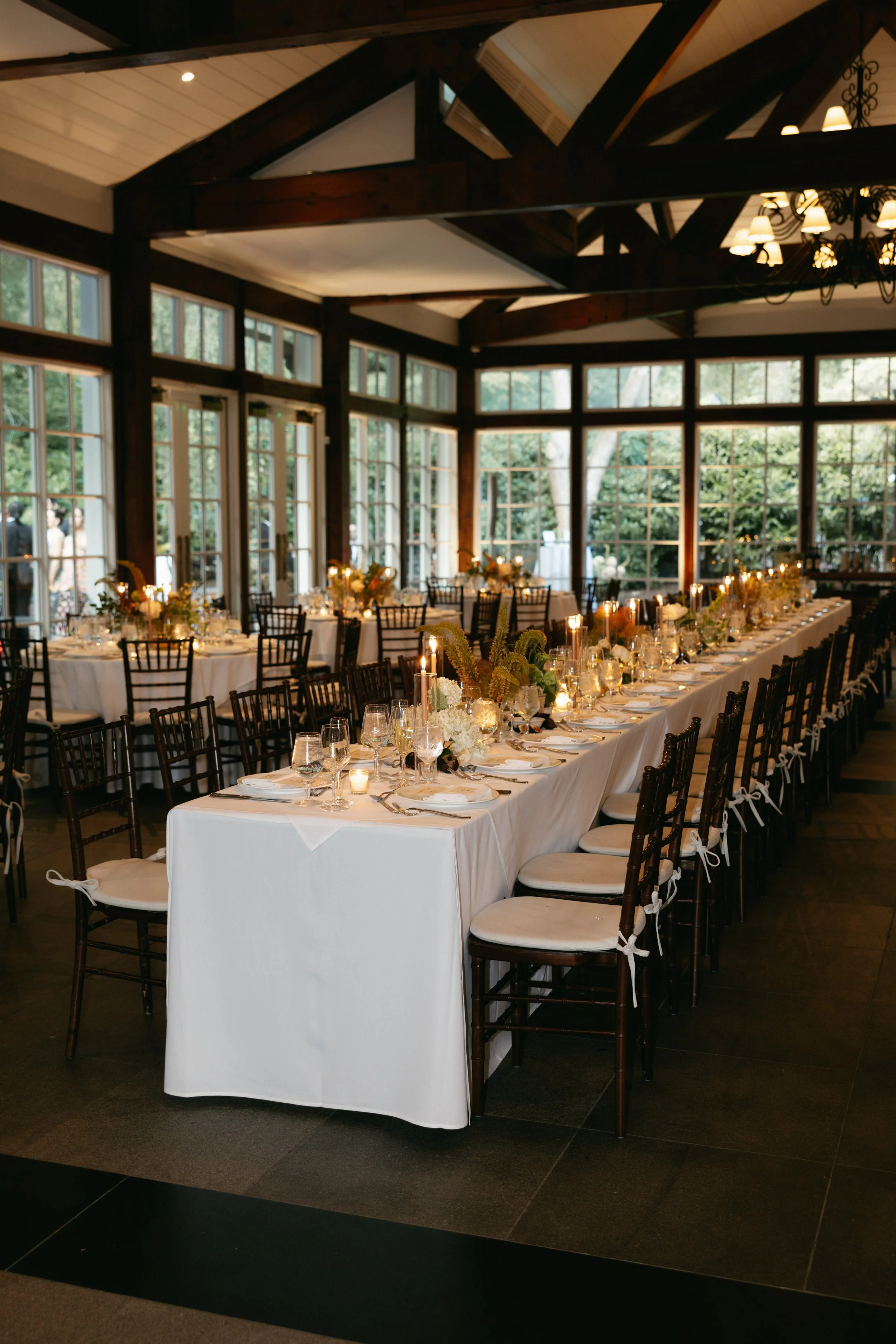 Long banquet table set with white tablecloths, candles, glassware, and floral centerpieces in a bright room with large windows and wooden beams. marigolds florals nyc florist wedding florals central park boathouse wedding