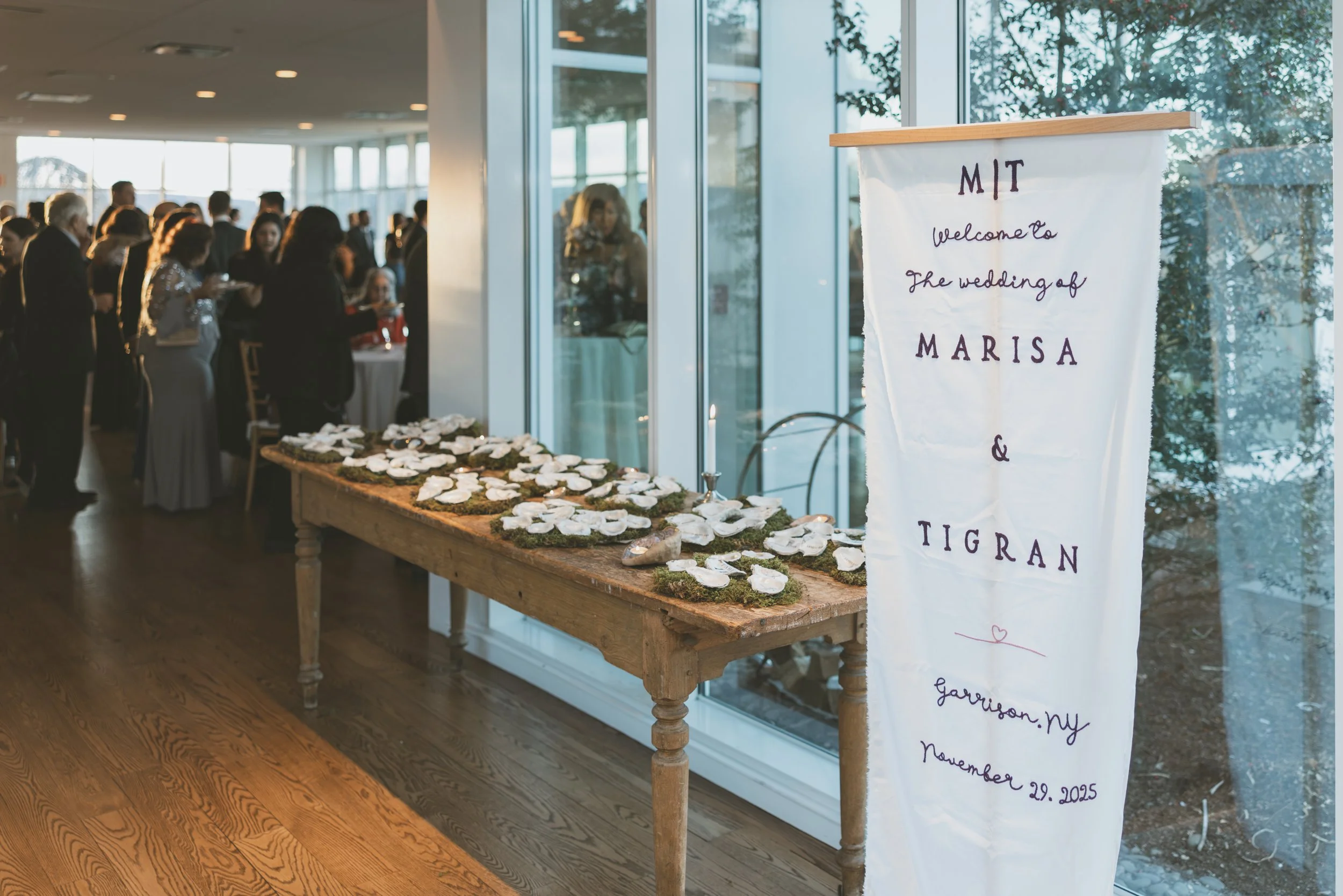 A reception area with a group of people gathered near a table decorated with small white items, possibly wedding favors or decorations. There is a large window on the right with trees visible outside. A white wedding sign with purple writing indicate