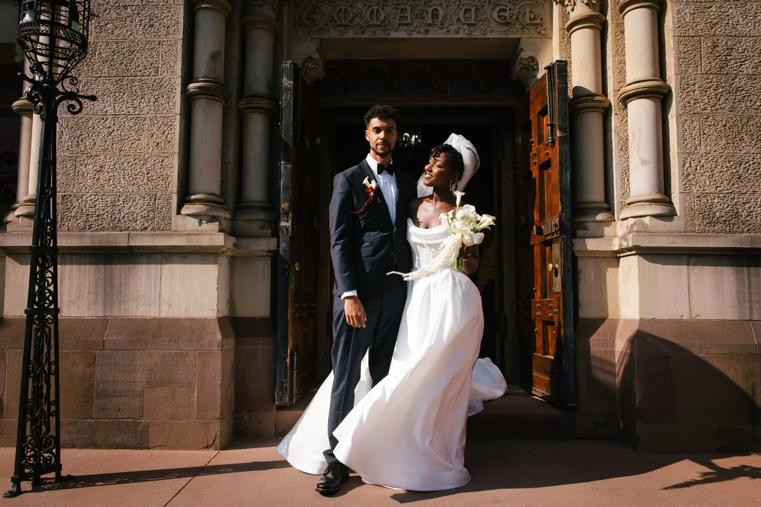 A newlywed couple standing at the entrance of a church on their wedding day, with the bride holding a bouquet of white flowers and the groom in a black tuxedo.