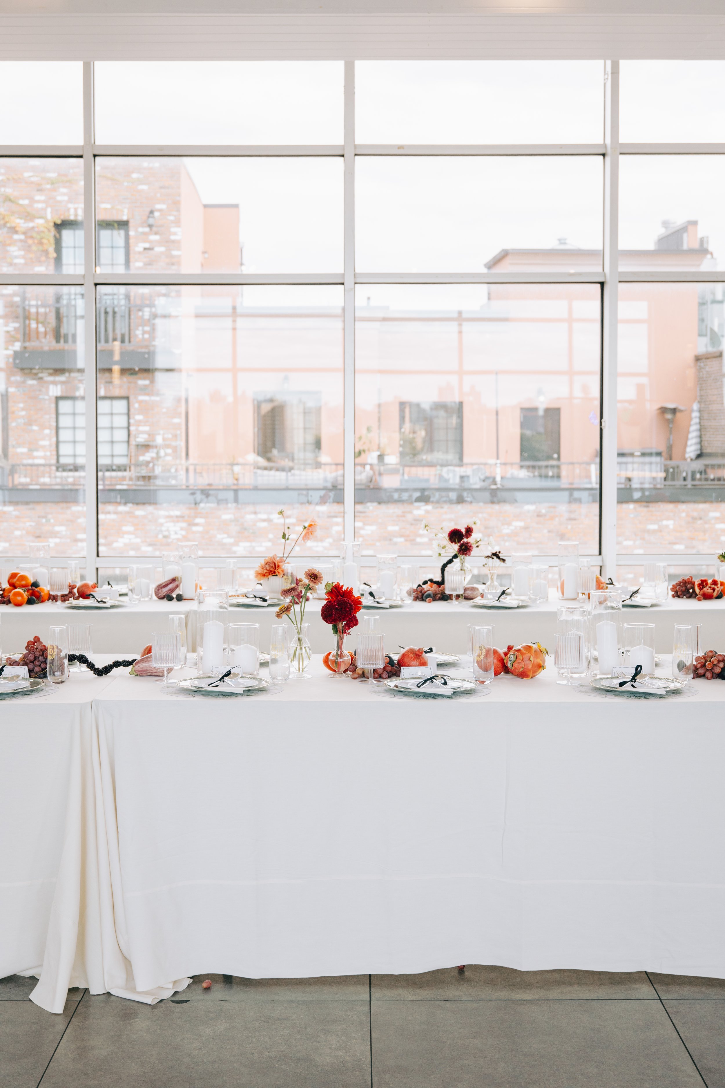 A long banquet table set with white tablecloths, glassware, and plates, decorated with flower arrangements and autumnal fruits, in front of a large window with a cityscape view. marigolds florals nyc florist wedding florals the w loft wedding