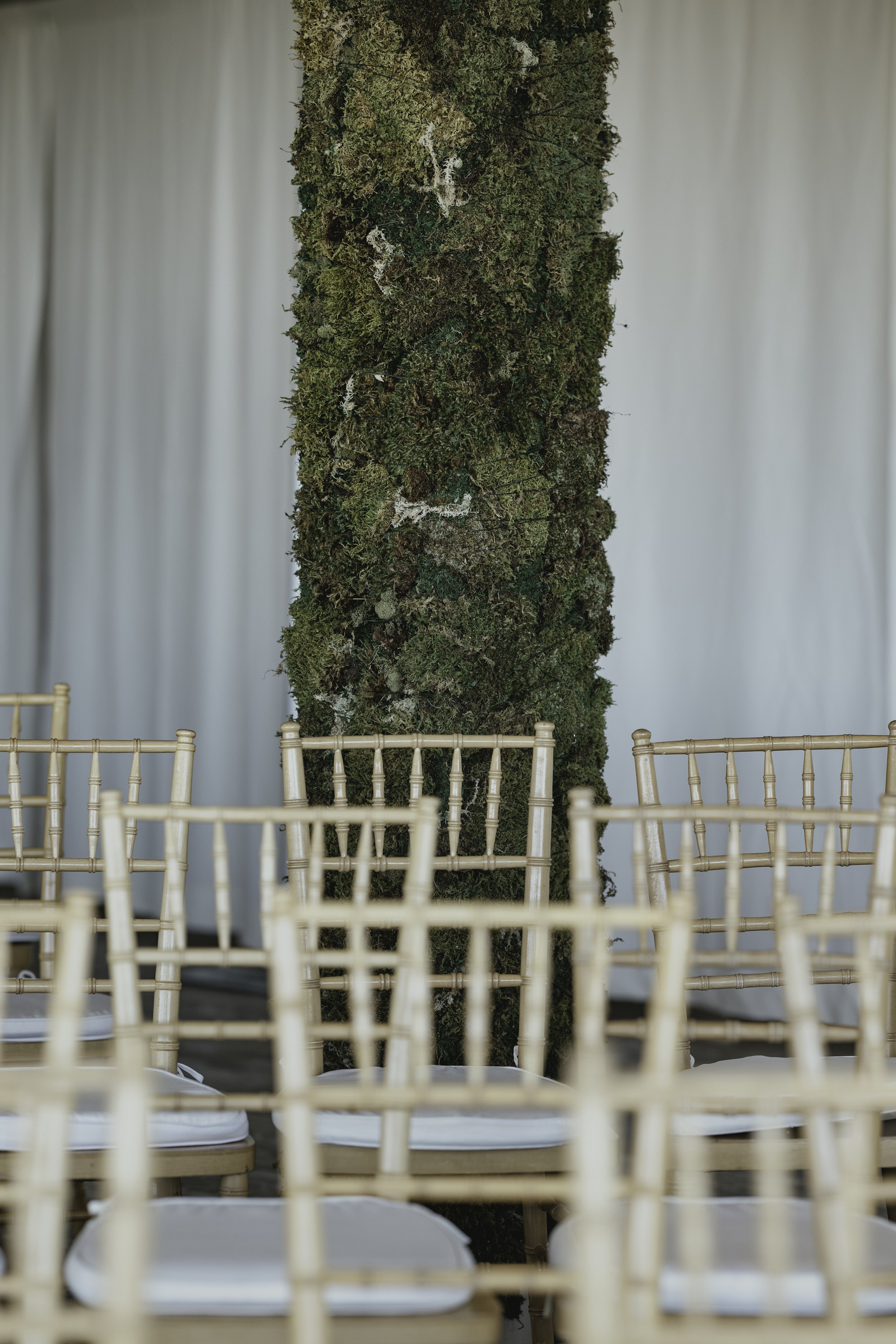 A large, vertical, moss-covered tree trunk standing in a room with a white curtain backdrop. Several empty chairs with white seats are arranged in front of the tree. marigolds florals nyc florist wedding florals the garrison