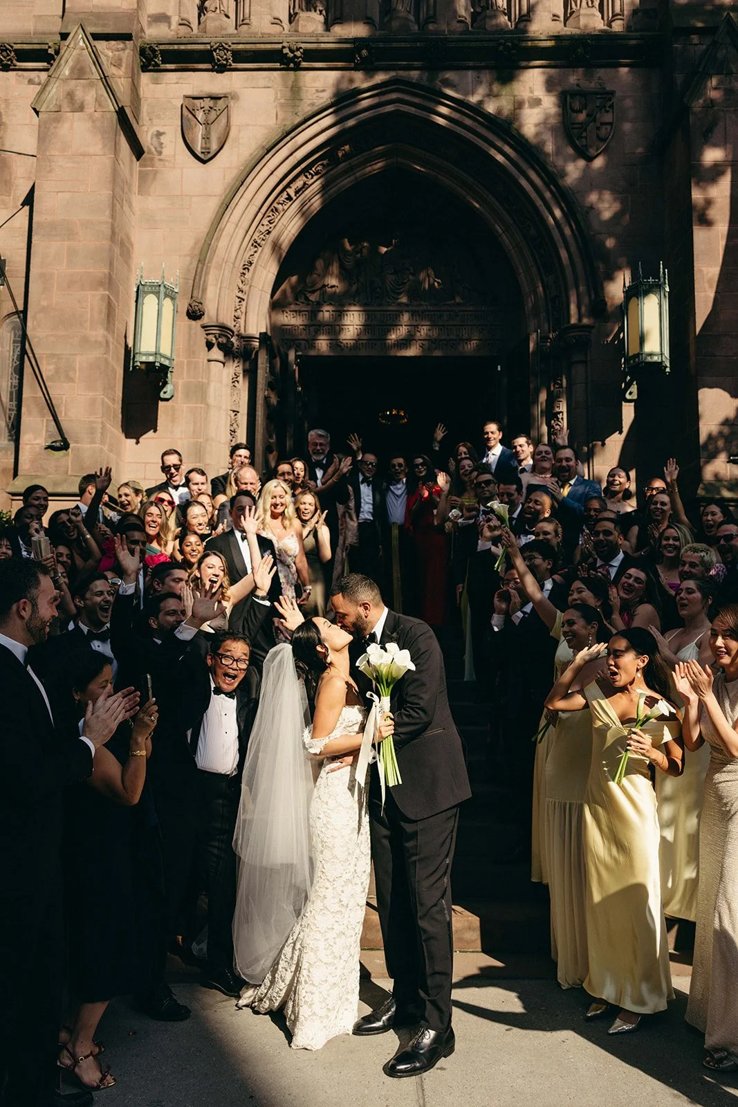 A wedding celebration scene outside a church with a bride and groom kissing, surrounded by many guests cheering and taking photos. marigolds florals nyc florist wedding florals central park boathouse wedding