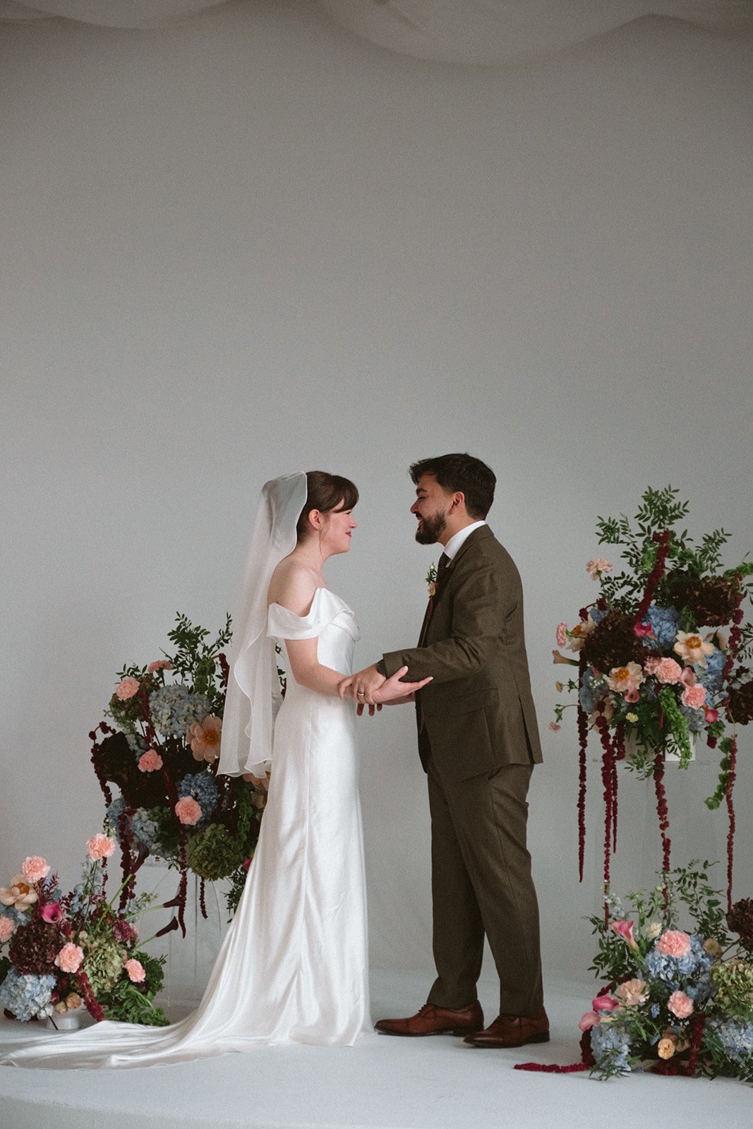 A bride and groom holding hands and smiling at each other during their wedding ceremony, surrounded by colorful floral arrangements. marigolds florals nyc florist wedding florals atmosfera wedding