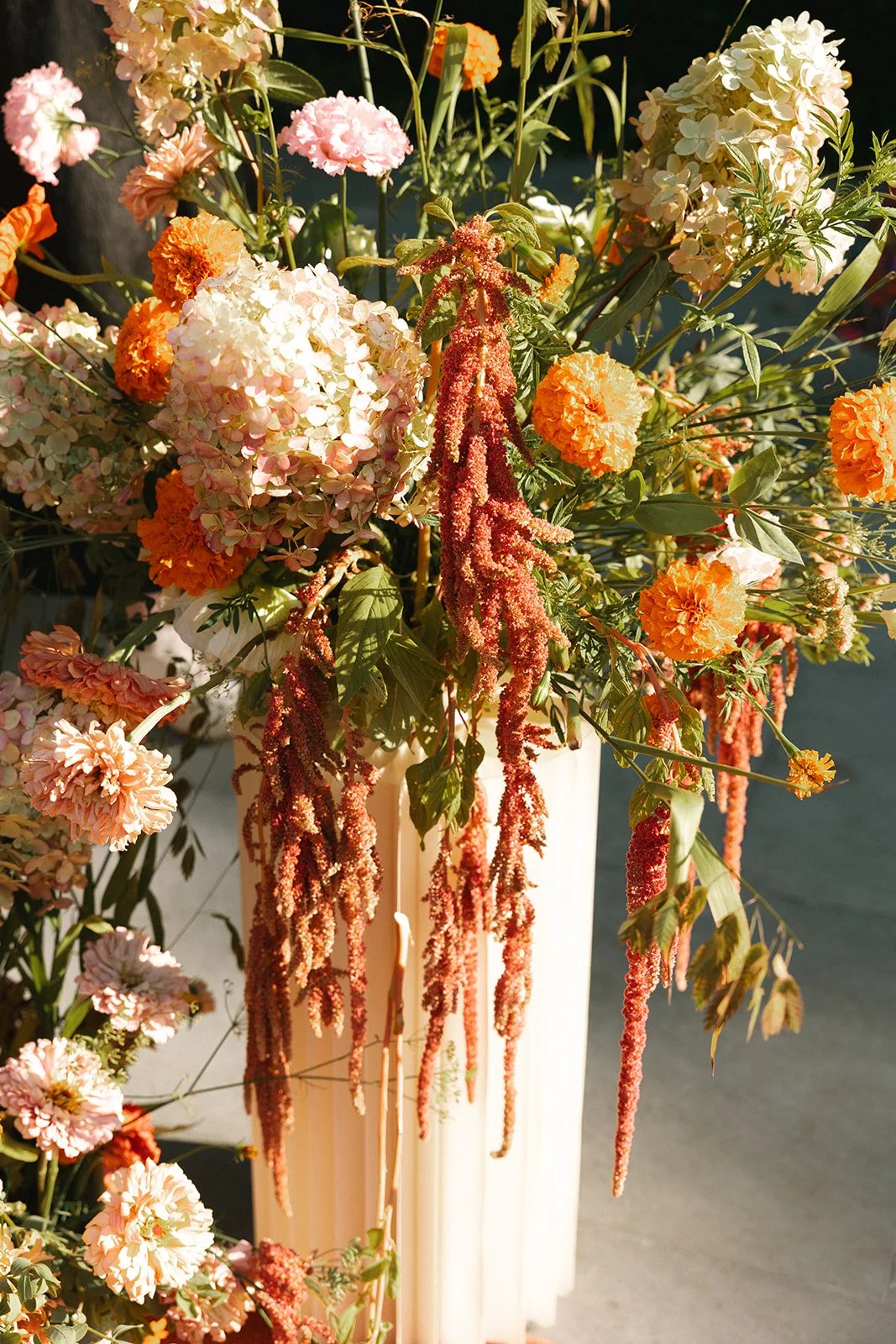 A bouquet of various flowers including pink and white hydrangeas, orange carnations, and hanging red amaranthus in a cream-colored vase. marigolds florals nyc florist wedding florals