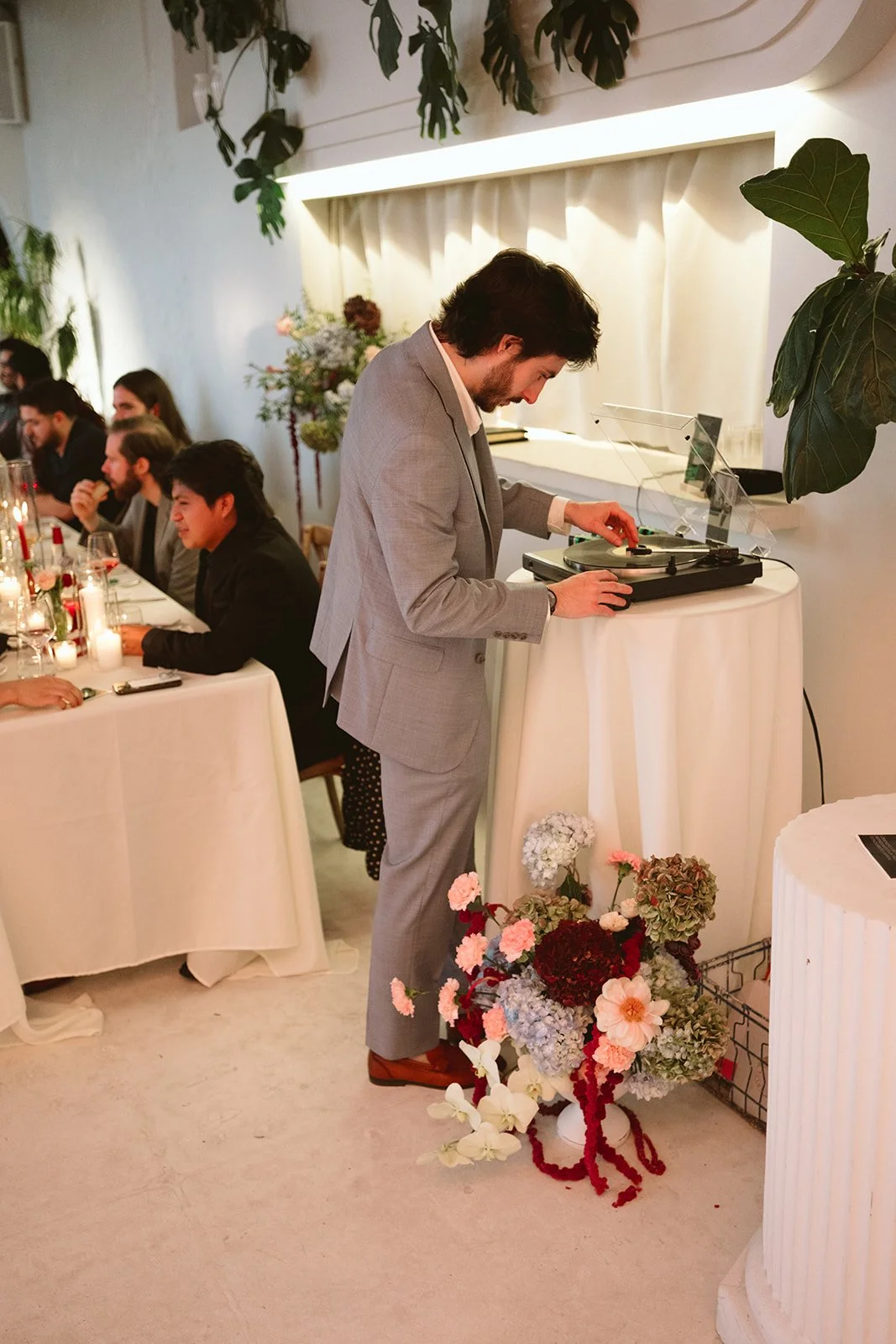 A man in a gray suit DJing at an indoor event with a table of seated guests, floral arrangements, and candles, with plants and a white wall in the background. marigolds florals nyc florist wedding florals atmosfera wedding intimate wedding