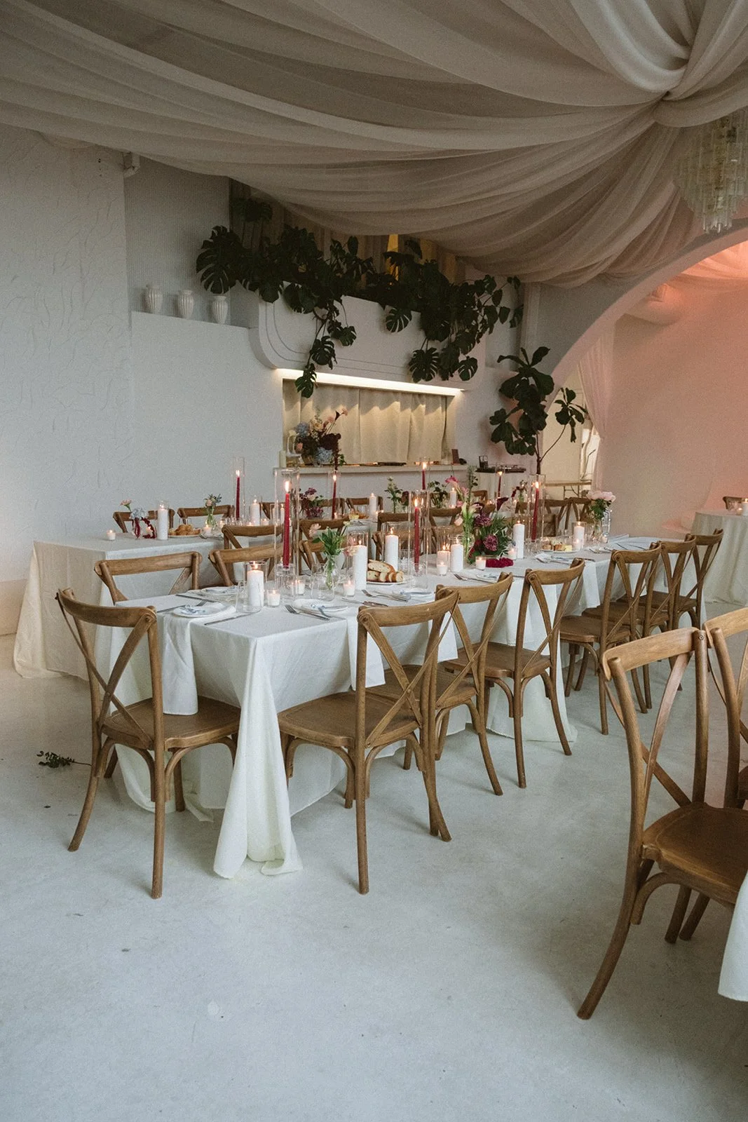 Elegant indoor wedding reception with a long table set with white tablecloths, candles, and floral centerpieces, featuring wooden chairs and lush green plants in a decorated room. marigolds florals nyc florist wedding florals atmosfera wedding