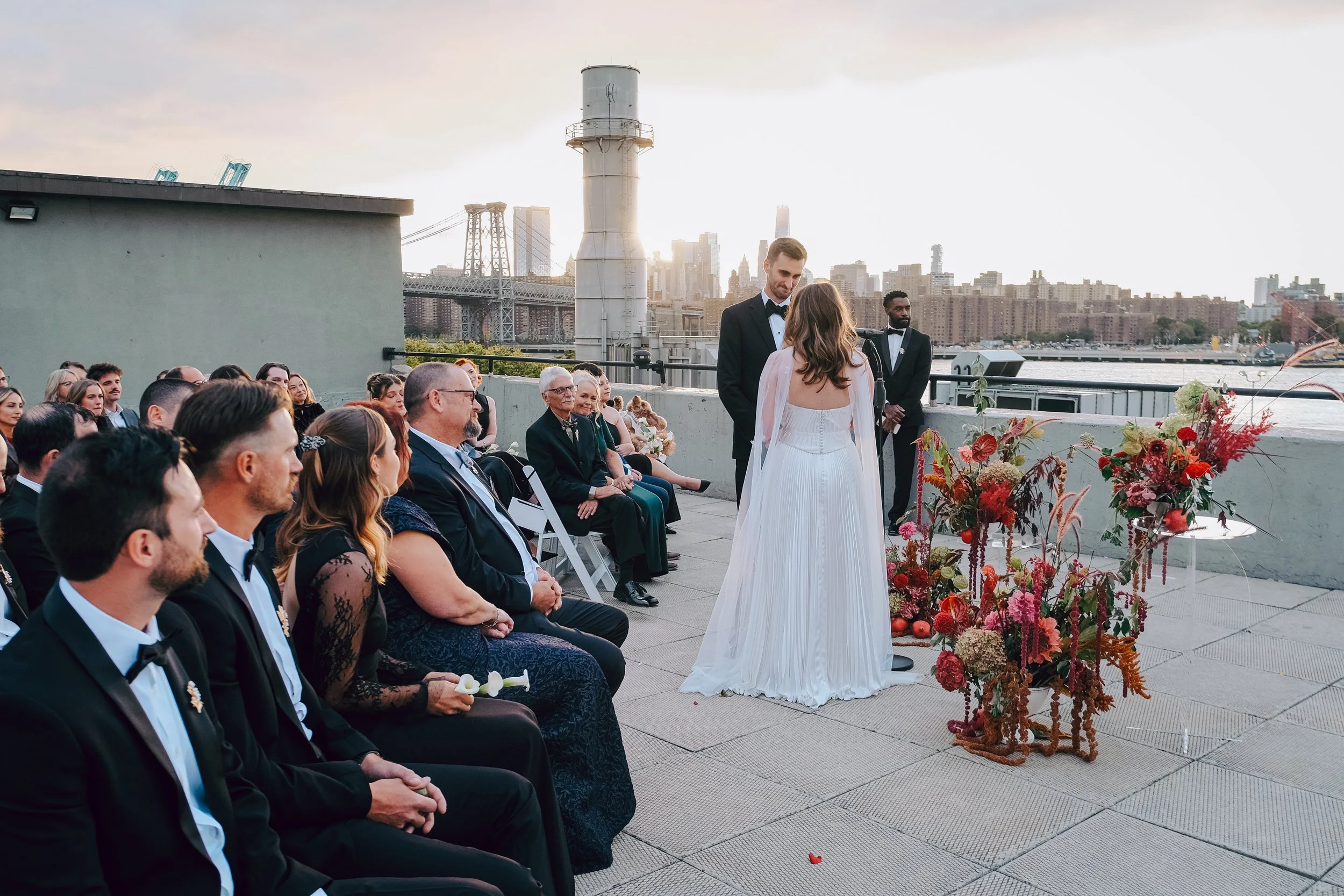 A wedding ceremony on a rooftop with city skyline, bridge, and river in the background. Bride and groom are standing facing each other, surrounded by seated guests. The bride is in a white dress, and the groom is in a tuxedo. A large floral arrangeme