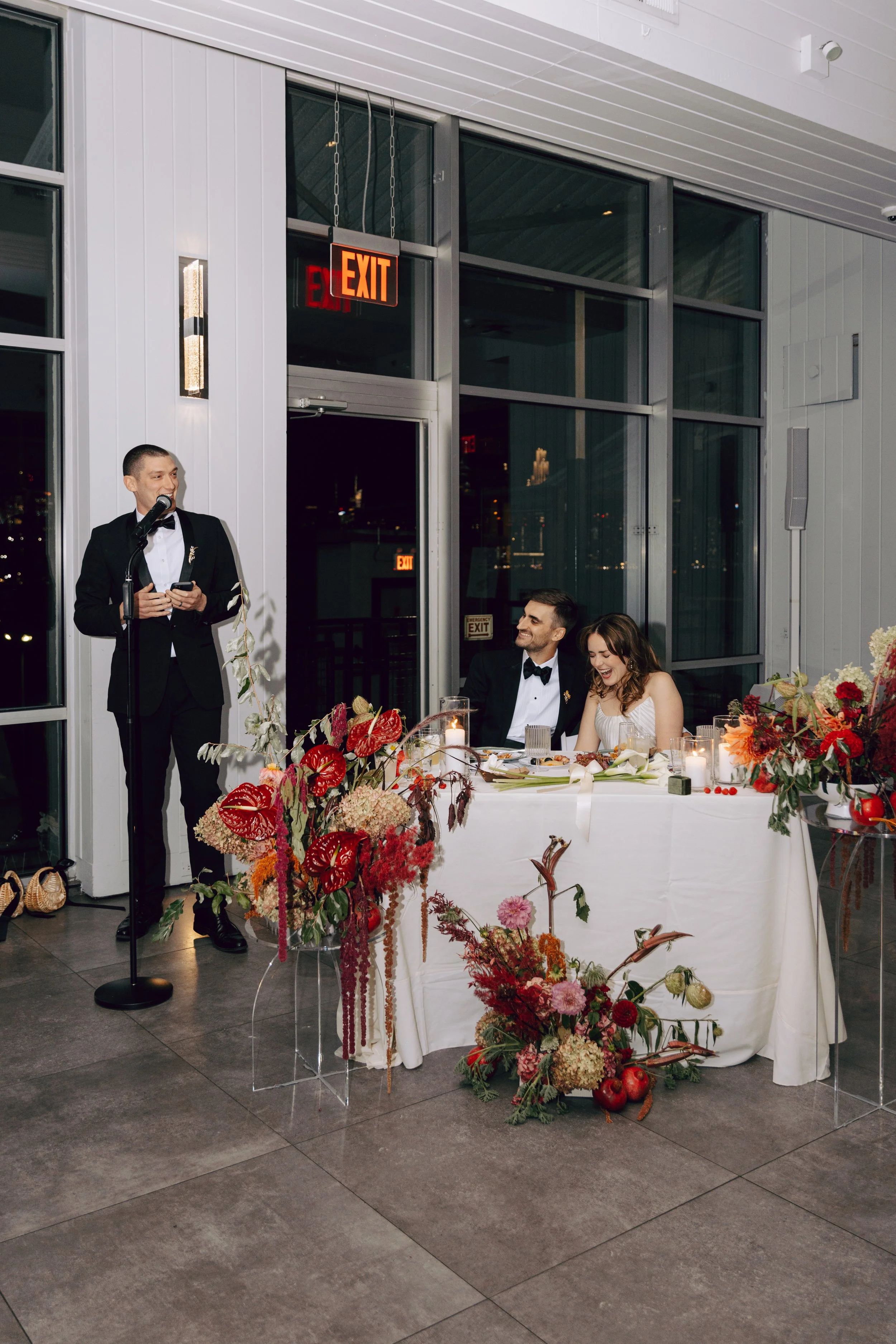 A wedding reception scene with a bride and groom seated at a decorated table, a man giving a speech at a microphone, and floral arrangements on the table and around the setting, inside a modern venue with large windows and closed exit signs.