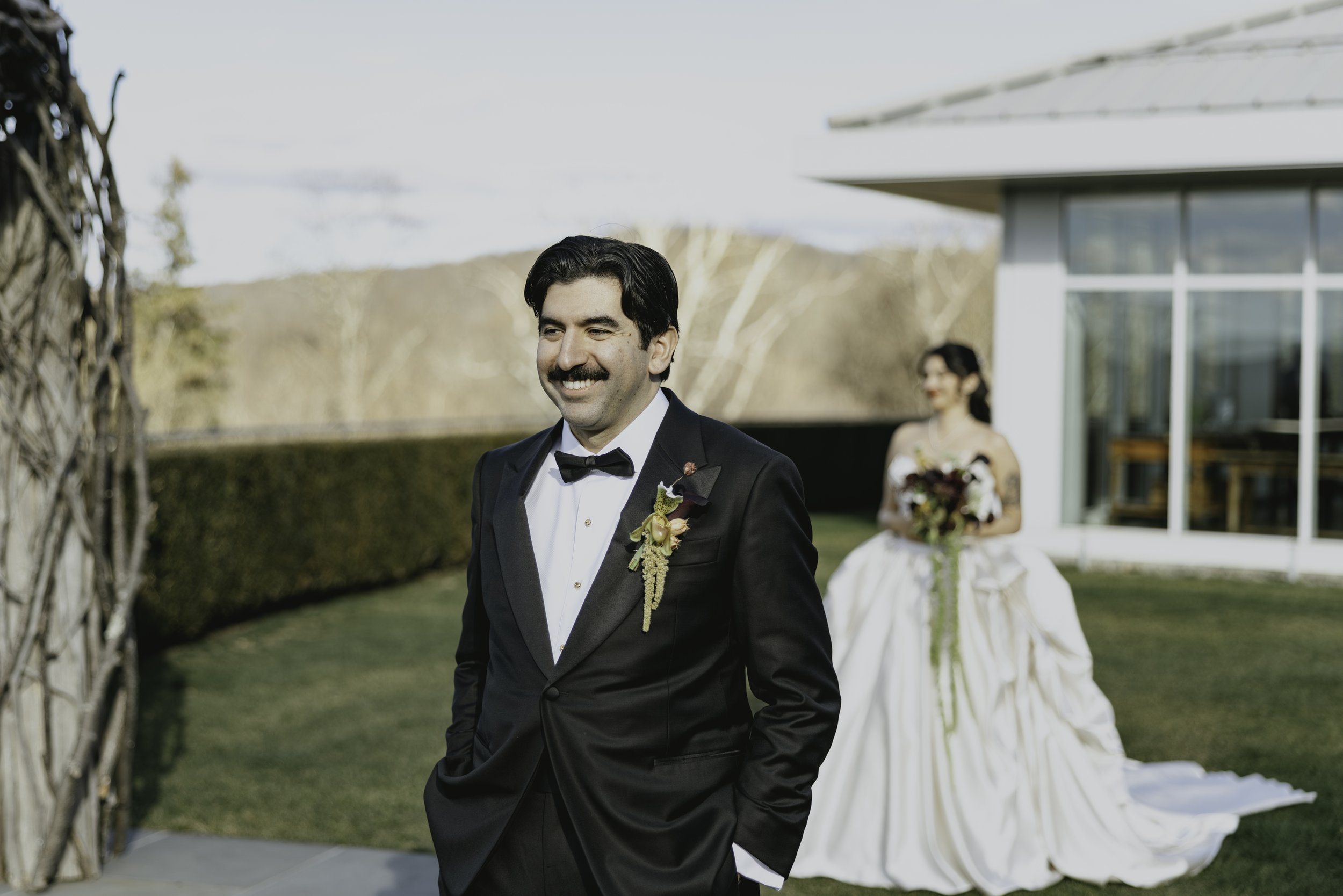 A man with dark hair and a mustache wearing a black tuxedo with a white shirt and black bow tie, smiling outdoors during a wedding, with a woman in a wedding dress holding a bouquet in the background. marigolds florals nyc florist wedding florals