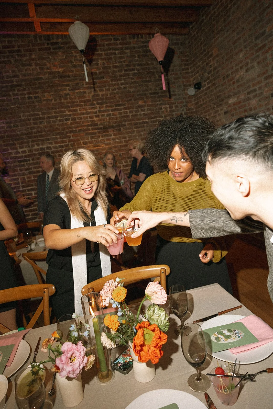 People at a party clinking glasses with drinks in a cozy brick-walled setting, with a table decorated with flowers and place settings. marigolds florals nyc florist wedding florals