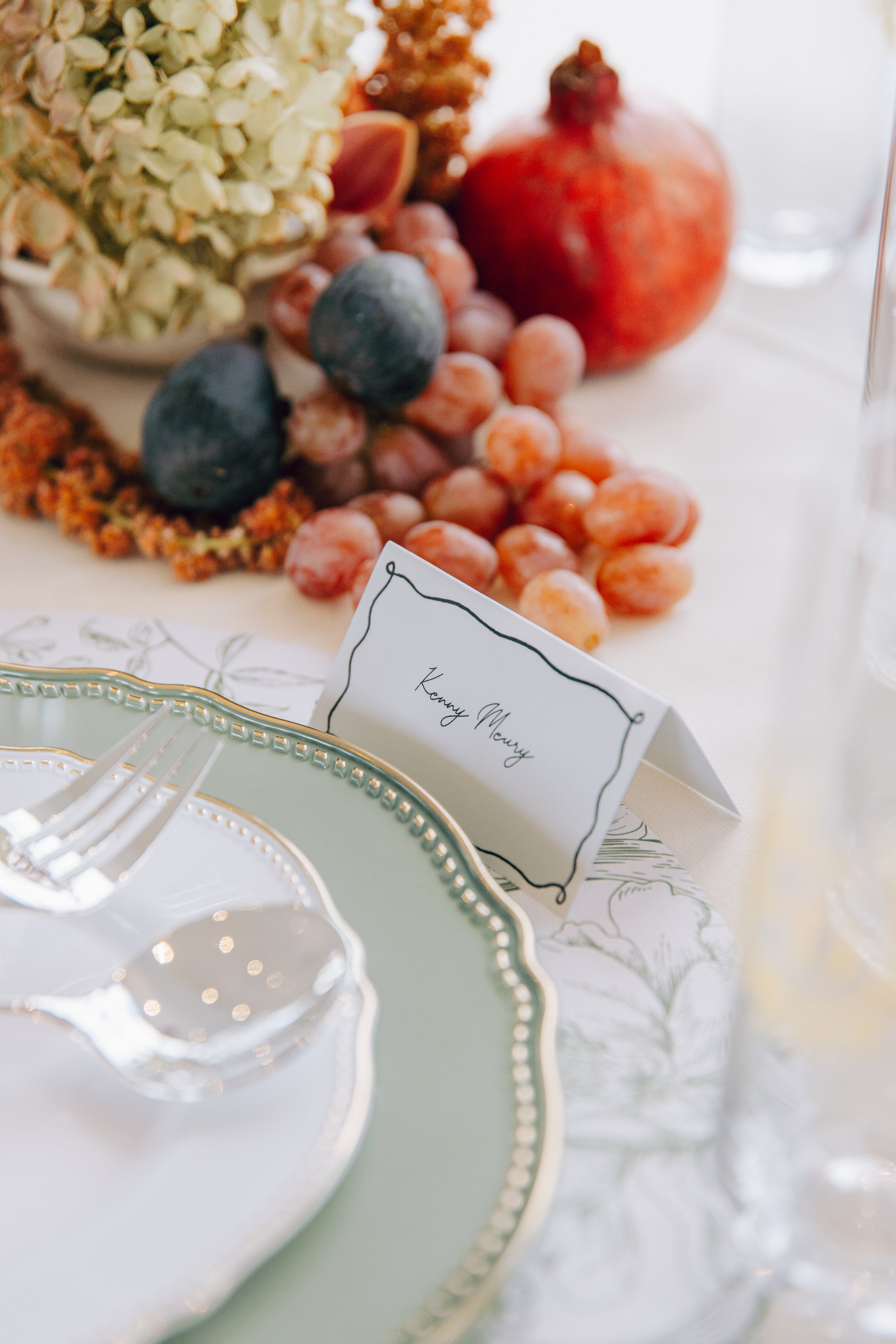 Close-up of a table setting featuring a white and gold decorative plate, with a display of grapes, pomegranate, and dried flowers in the background. marigolds florals nyc florist wedding florals the w loft wedding pedestal wedding ceremony anthurium 