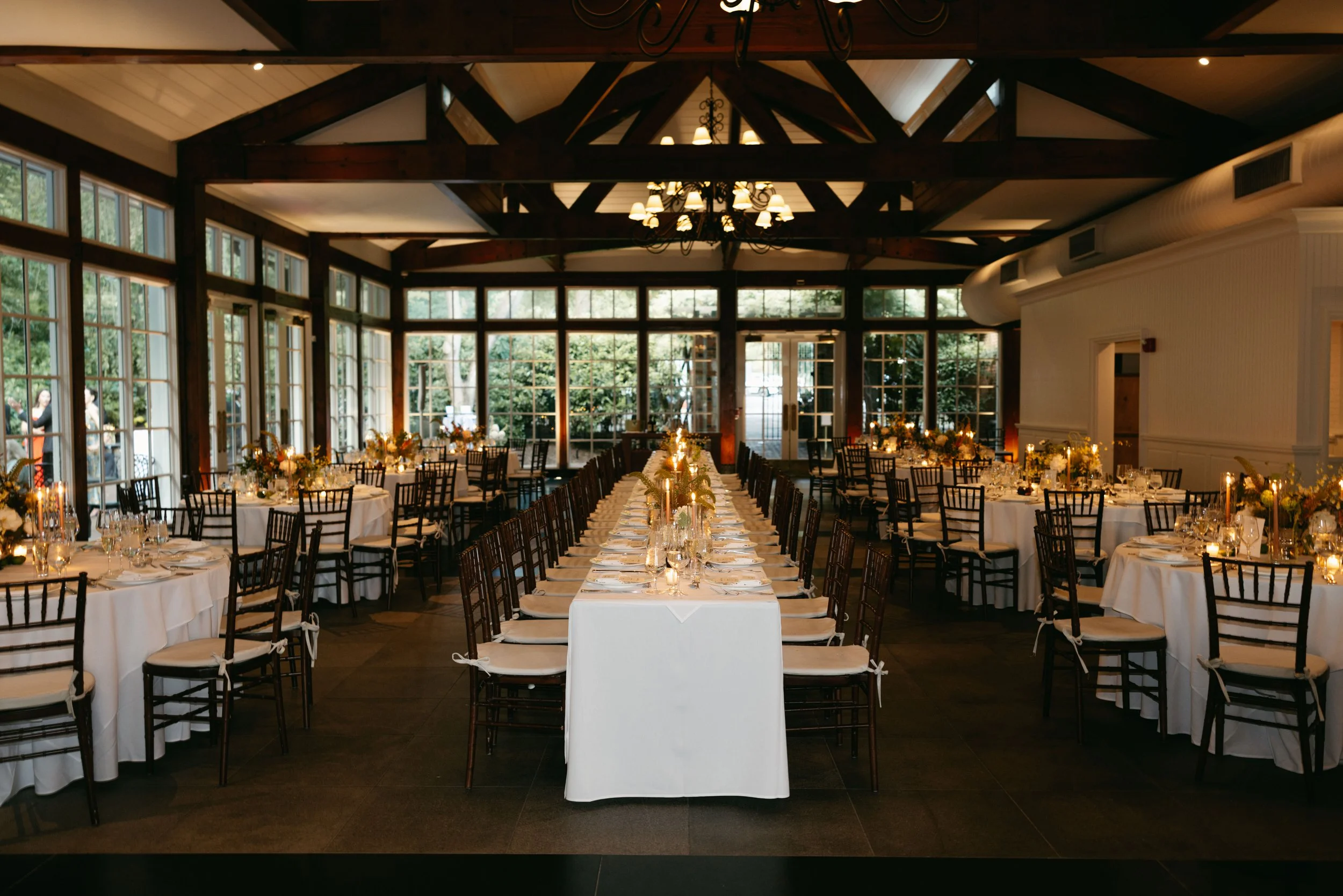 Elegant banquet hall with a long center table decorated with flowers and candles, surrounded by chairs with white cushions, large windows, and a chandelier hanging from a high wooden-beamed ceiling.