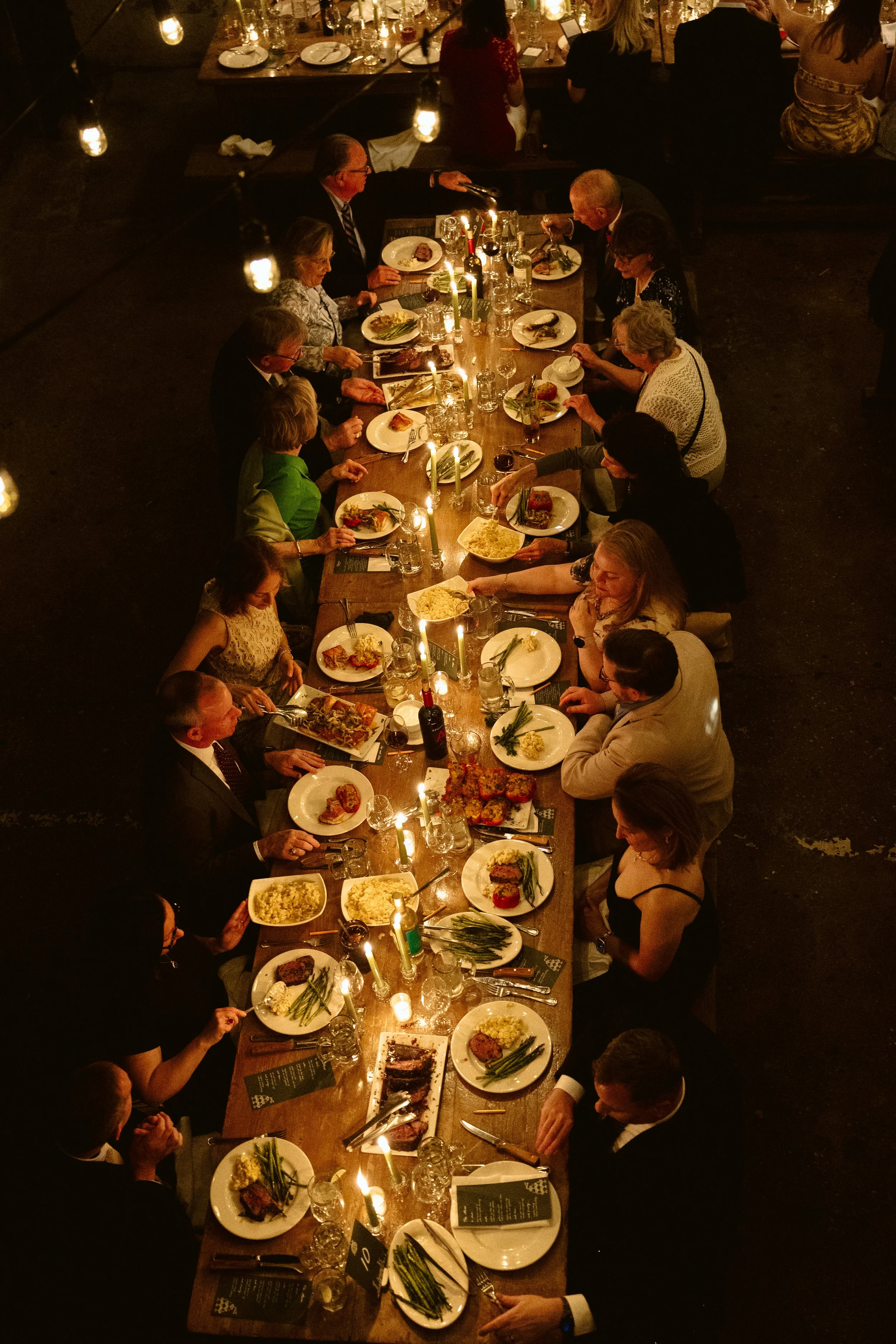 A long dining table is set for a formal dinner with multiple guests. The table has plates of food, glasses, candles, and wine bottles. The scene is dimly lit, creating an intimate atmosphere, with people engaged in conversation and enjoying their mea