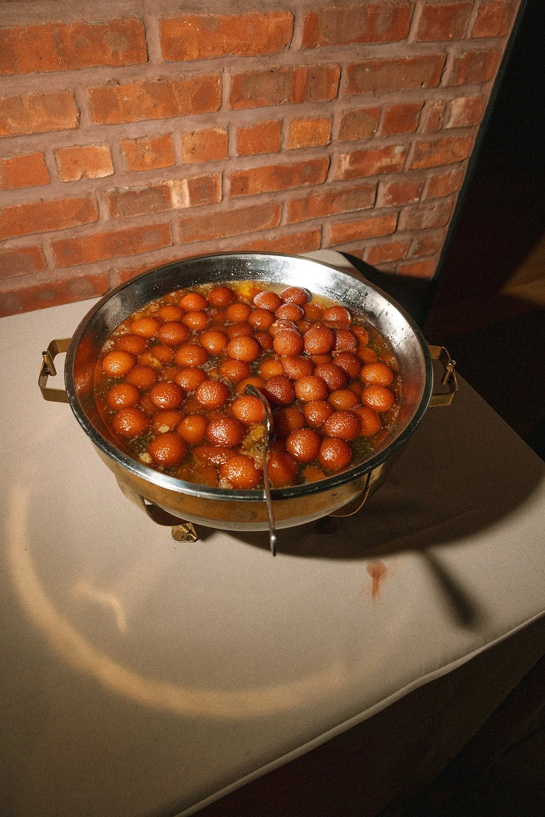 A metal tray with small round, golden-brown fried pastries or balls floating in syrup, placed on a white table against a brick wall background. marigolds florals nyc florist wedding florals
