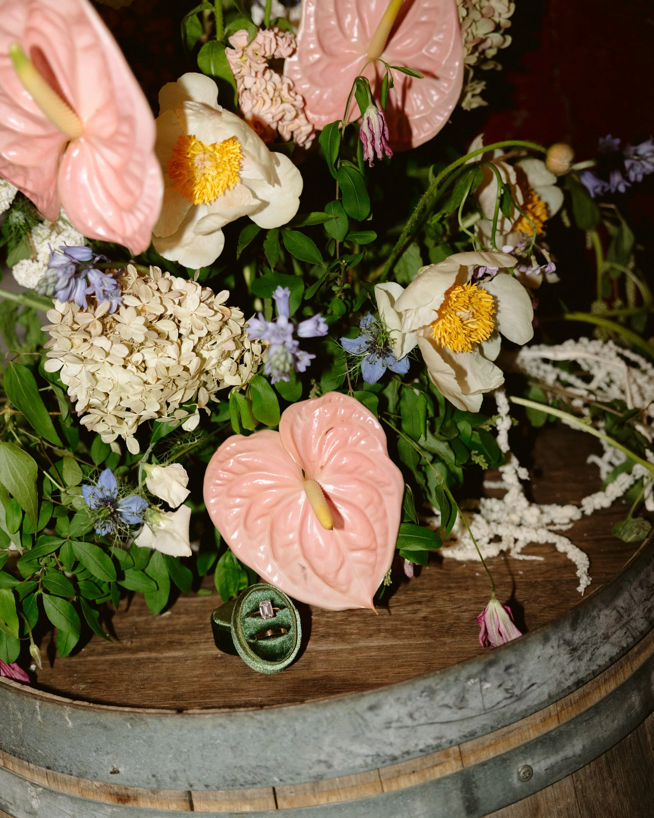A flower arrangement with pink anthuriums, white poppies, hydrangea, and other small flowers in a wooden container, decorated with a green ring and a small jewelry box, on a wooden surface. marigolds florals nyc florist wedding florals
