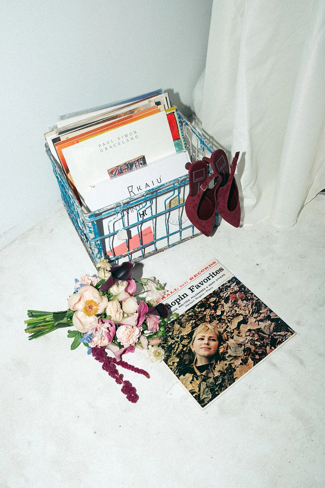 A blue wire basket holding magazines and papers, a pair of burgundy high-heeled shoes hanging on the side of the basket, a bouquet of pink and white flowers on the floor, and a magazine featuring a woman on the cover laid on a white surface marigolds