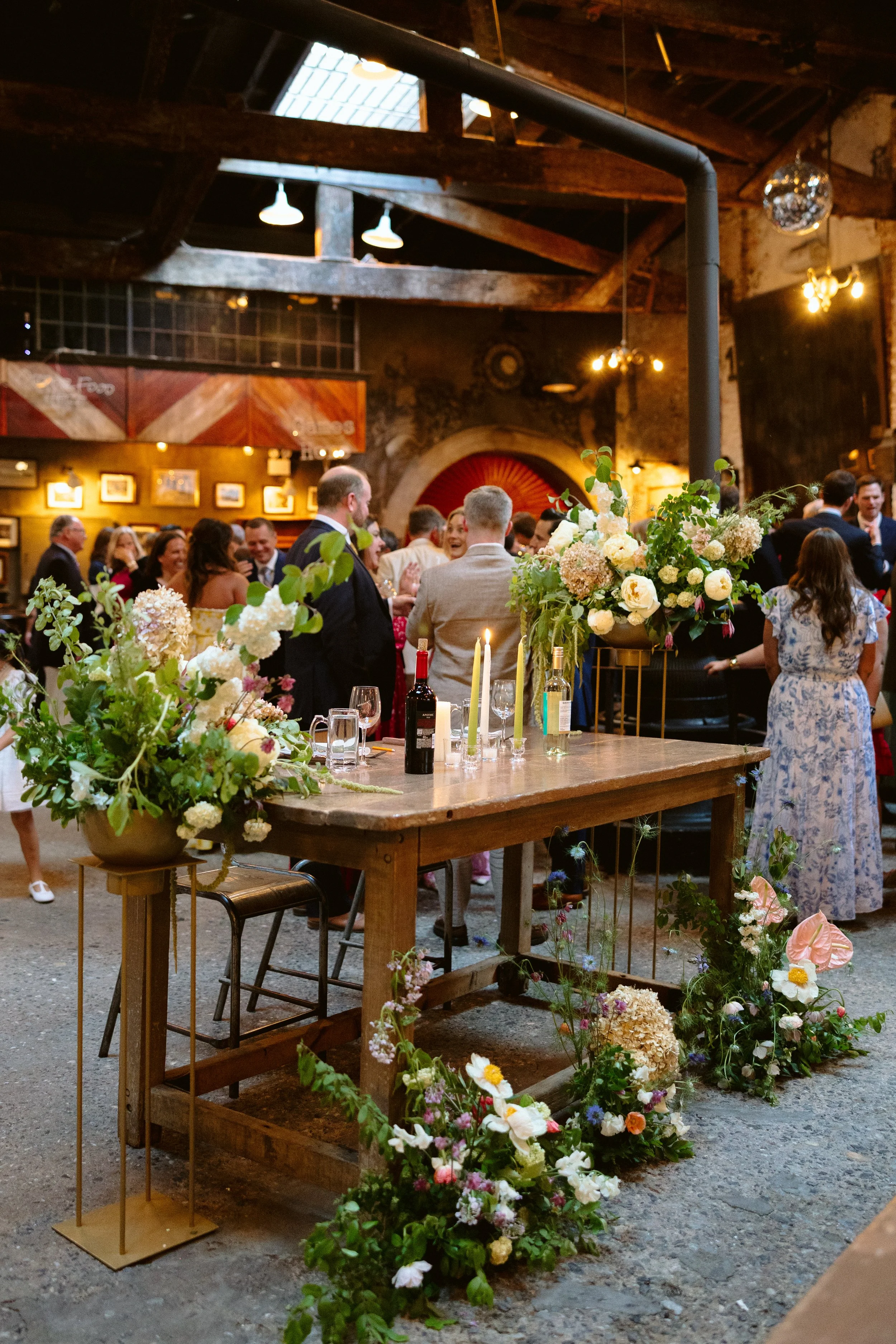 A lively wedding event with people dressed in formal attire,  tables decorated with elegant floral arrangements. The setting has a rustic charm with exposed brick walls, wooden beams, and ambient lighting marigolds florals nyc florist wedding florals