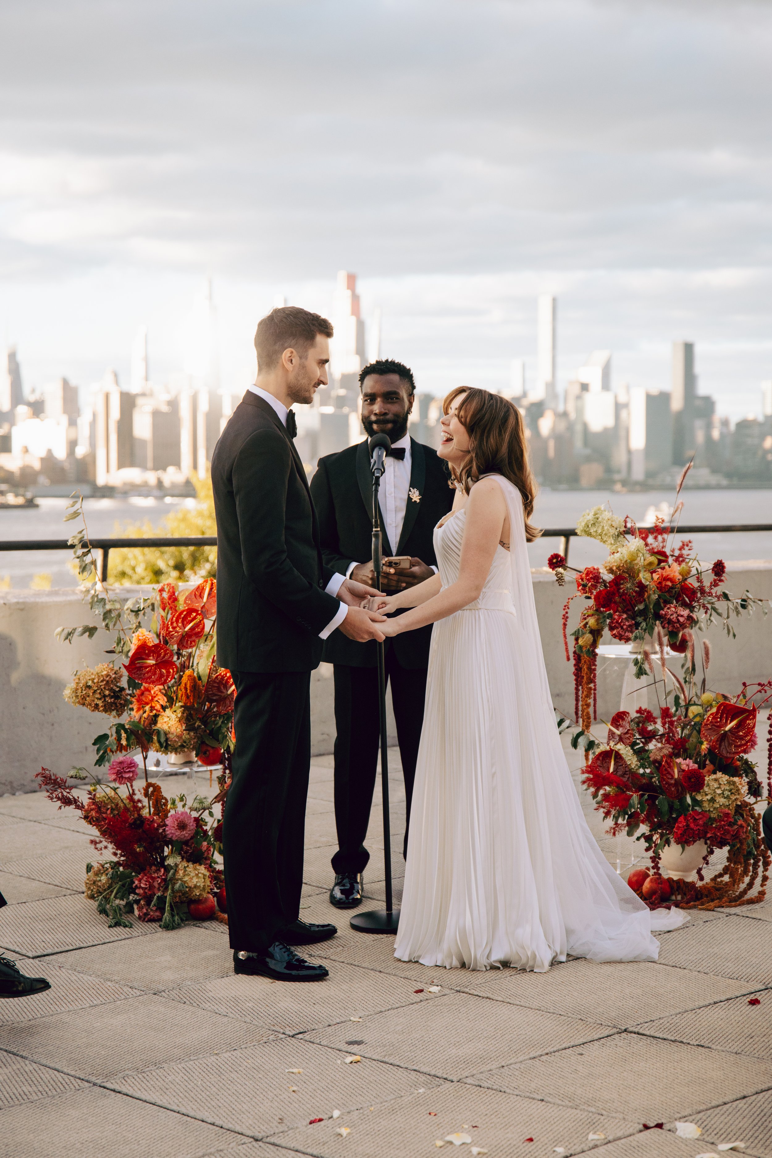 A couple getting married on a rooftop with city skyline in the background. They are holding hands, facing each other, with an officiant standing behind them, smiling. The bride is wearing a white wedding dress, and the groom is in a black tuxedo. The