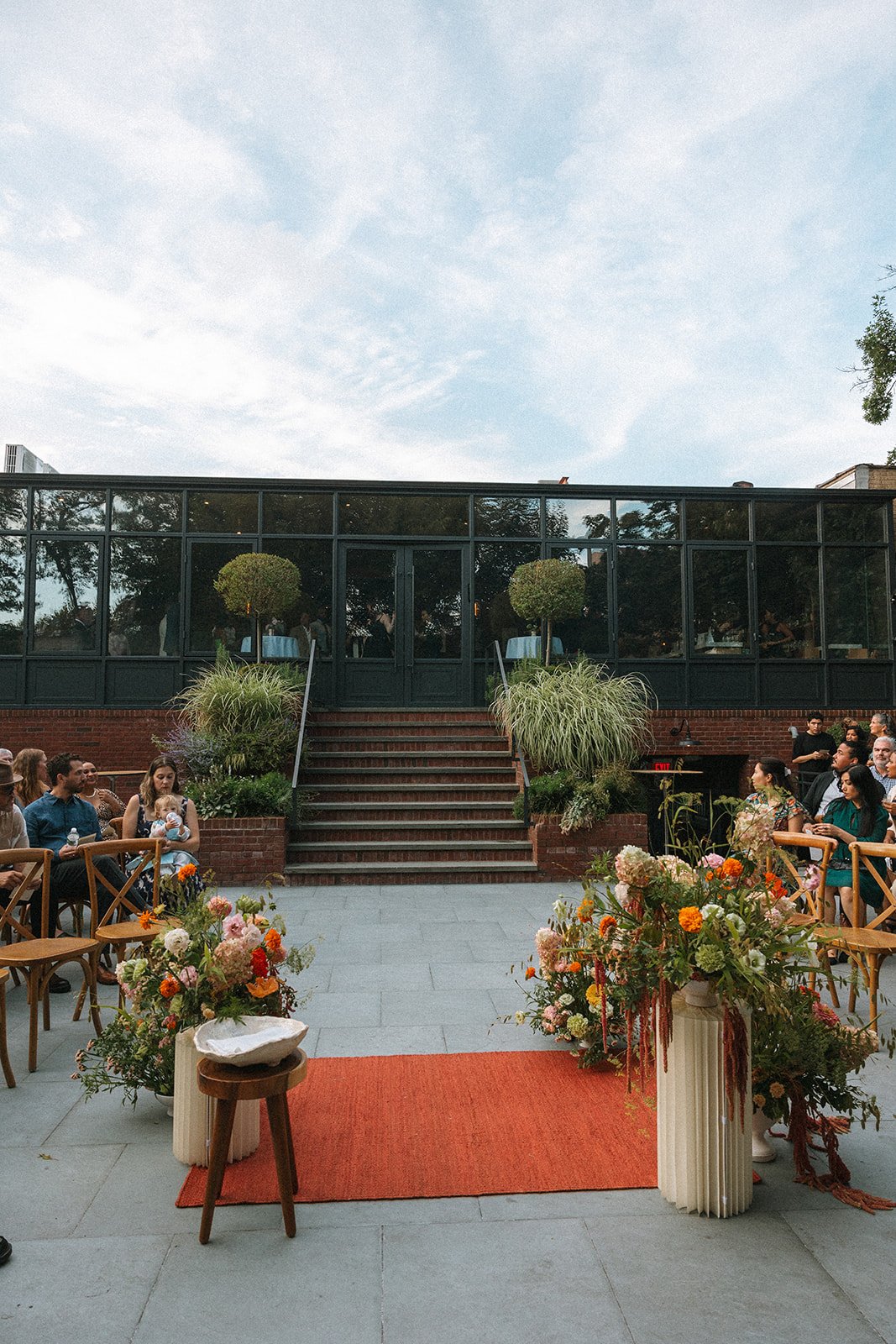 Outdoor wedding ceremony setup with floral arrangements, chairs, and a brick building with large windows in the background. marigolds florals
