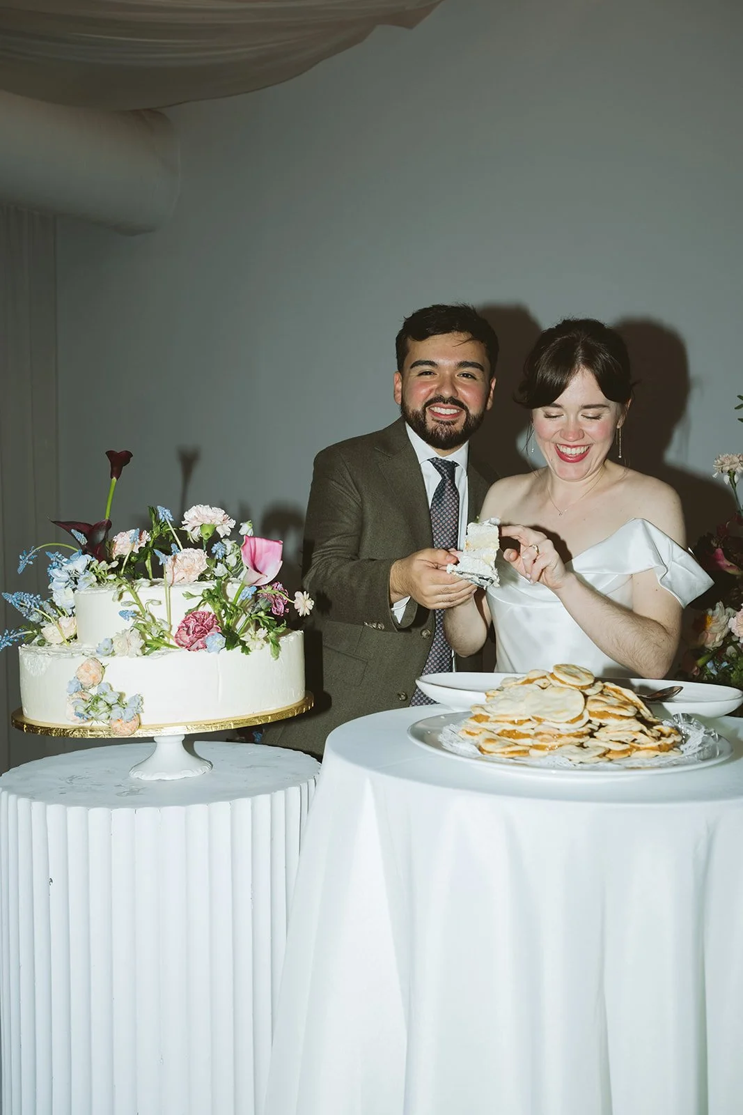 A couple at their wedding, cutting the wedding cake together, with a large floral decorated cake on a pedestal and a table of cookies in front of them, smiling and celebrating. marigolds florals nyc florist wedding florals atmosfera wedding