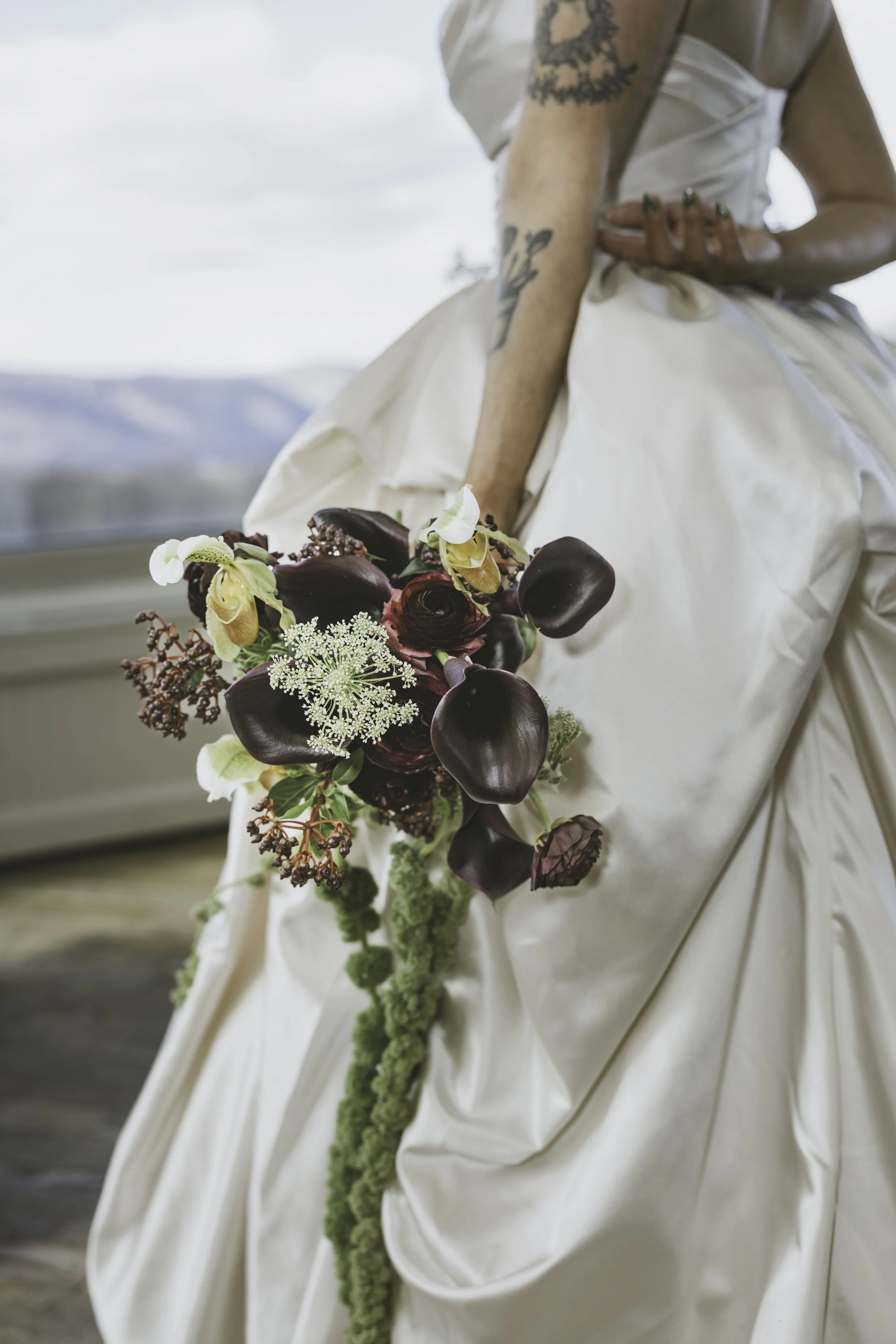 A person in a wedding dress holding a cascading bouquet of dark red, yellow, white, and green flowers. The person has tattoos on their arm, including a map outline, and has painted nails. The background shows a mountain landscape with a partly cloudy
