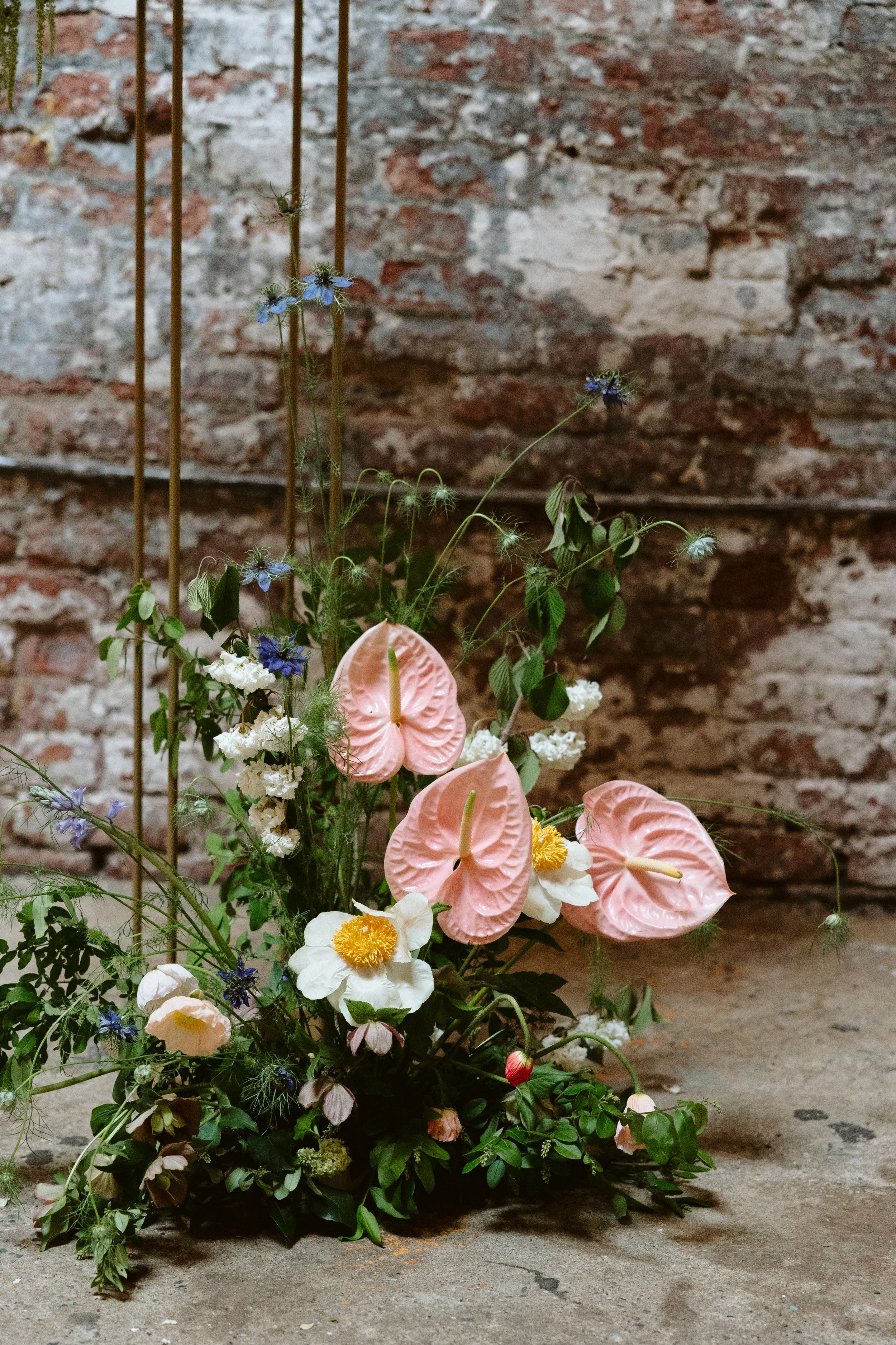 A floral arrangement with pink anthuriums, white flowers, and greenery placed on a concrete floor against a brick wall. marigolds florals nyc florist wedding florals houston hall nyc bar wedding