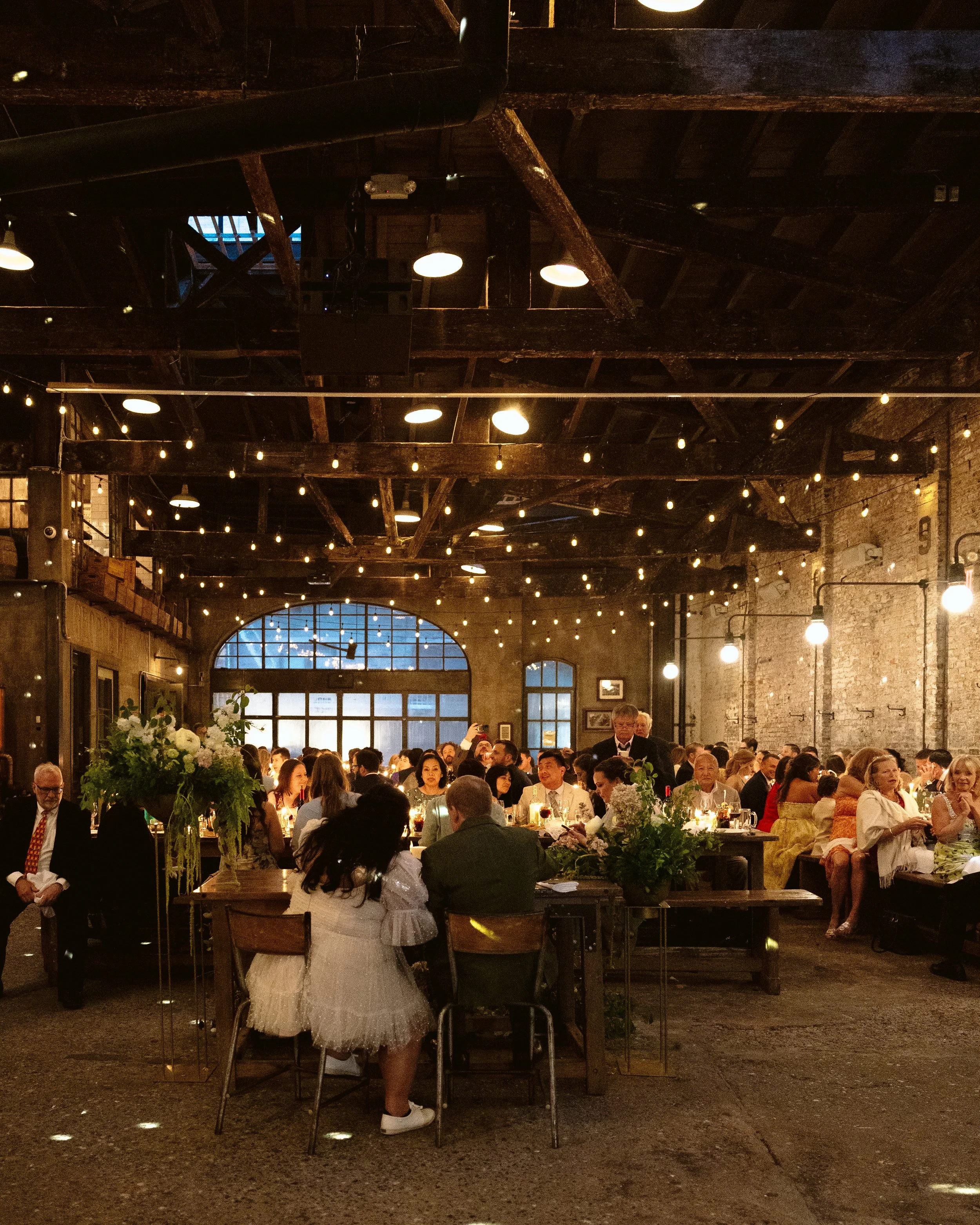 A lively indoor event with many people seated at tables decorated with flowers and candles, in a rustic space with exposed brick walls, large windows, string lights, and wooden beams. marigolds florals nyc florist wedding florals houston hall nyc bar