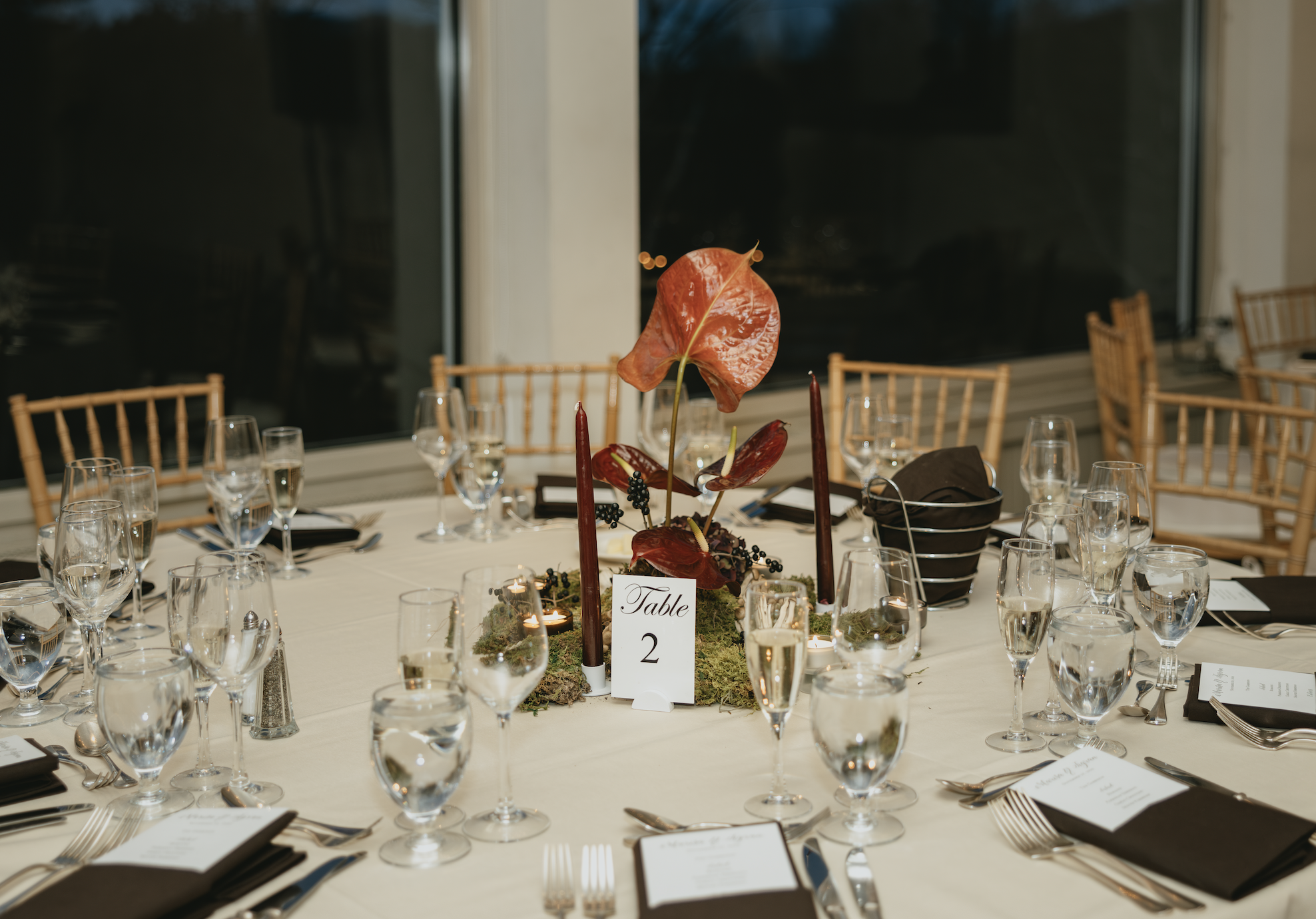 Elegant banquet table set for a formal event with glassware, folded black napkins, silverware, and a centerpiece with orange and red leaves, black candles, and a table number sign reading 'Table 2', next to windows at night.