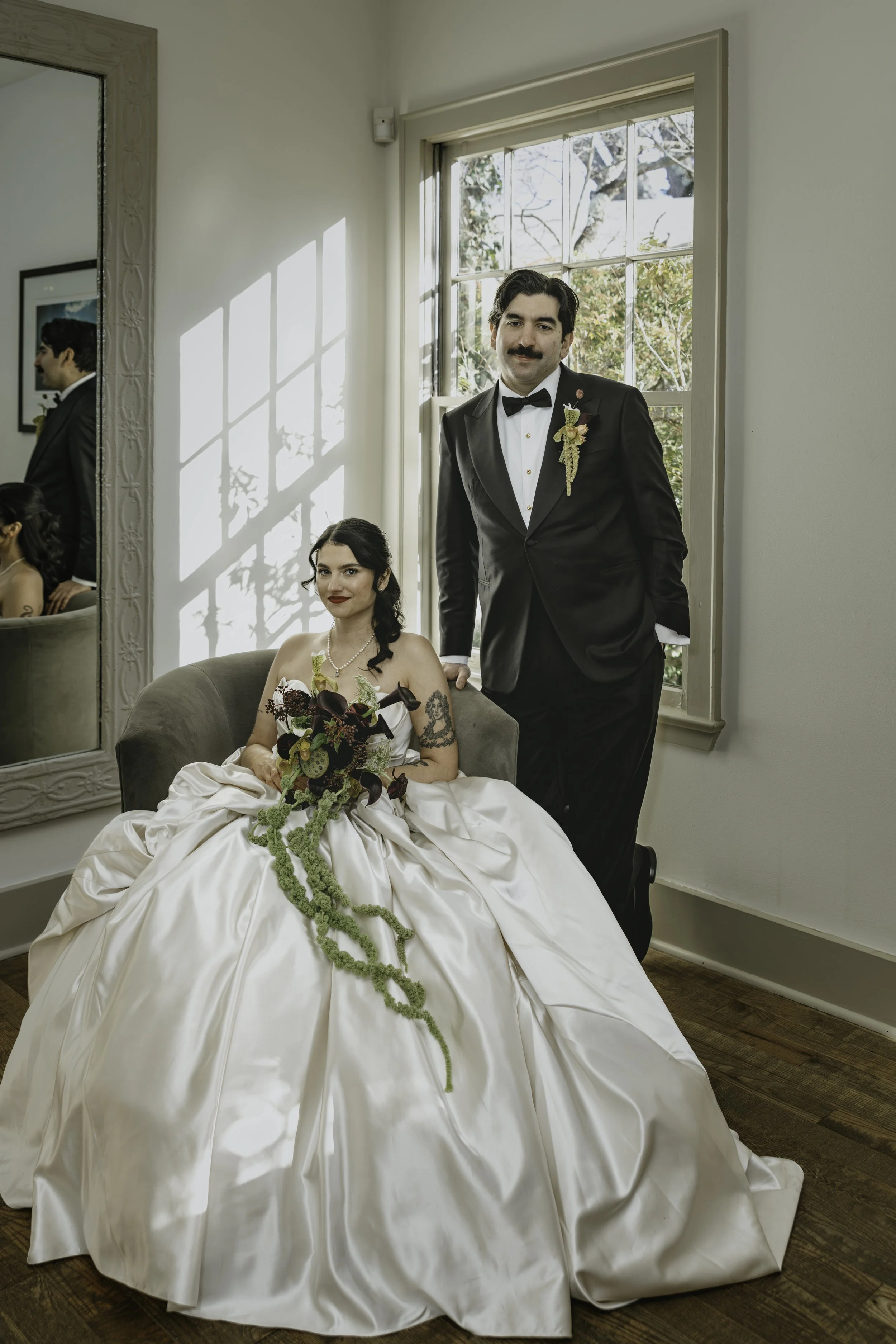 A man in a tuxedo with a boutonniere and a woman in a wedding dress holding a bouquet, sitting and standing near a window in a room with light-colored walls and a wooden floor.  marigolds florals nyc florist wedding florals the garrison