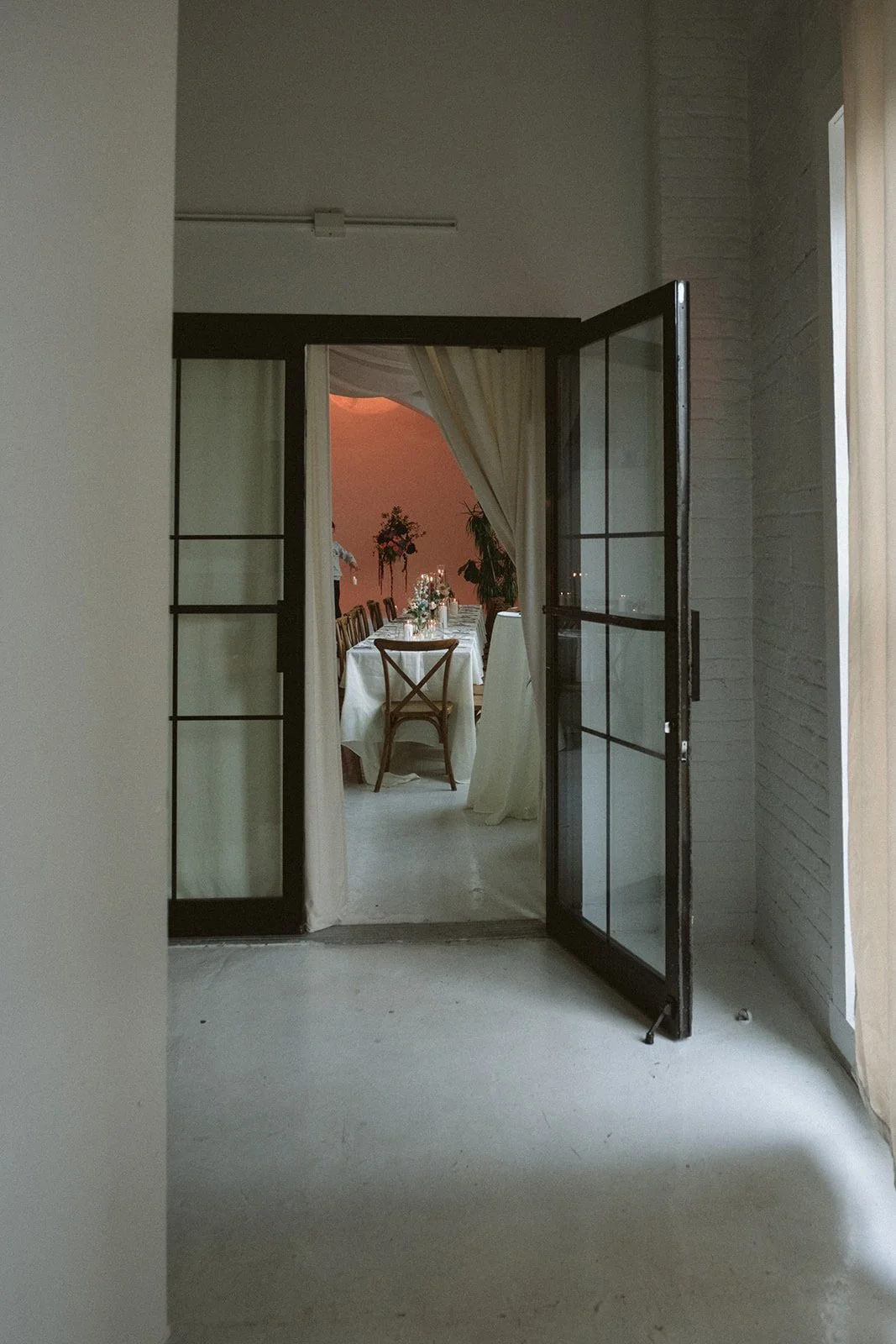 Open black-framed glass door revealing a dining room with a long table, chairs, floral centerpiece, and potted plants, with a pink sunset sky visible through the window. marigolds florals nyc florist wedding florals atmosfera wedding intimate wedding