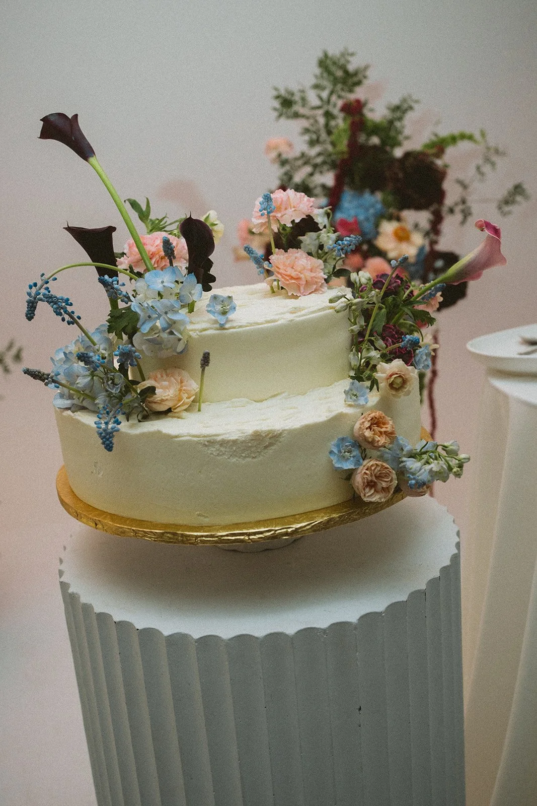 A two-tiered white wedding cake decorated with a variety of fresh pastel-colored flowers and dark calla lilies, placed on a gold cake board and sitting on a round white cake stand. marigolds florals nyc florist wedding florals atmosfera wedding 