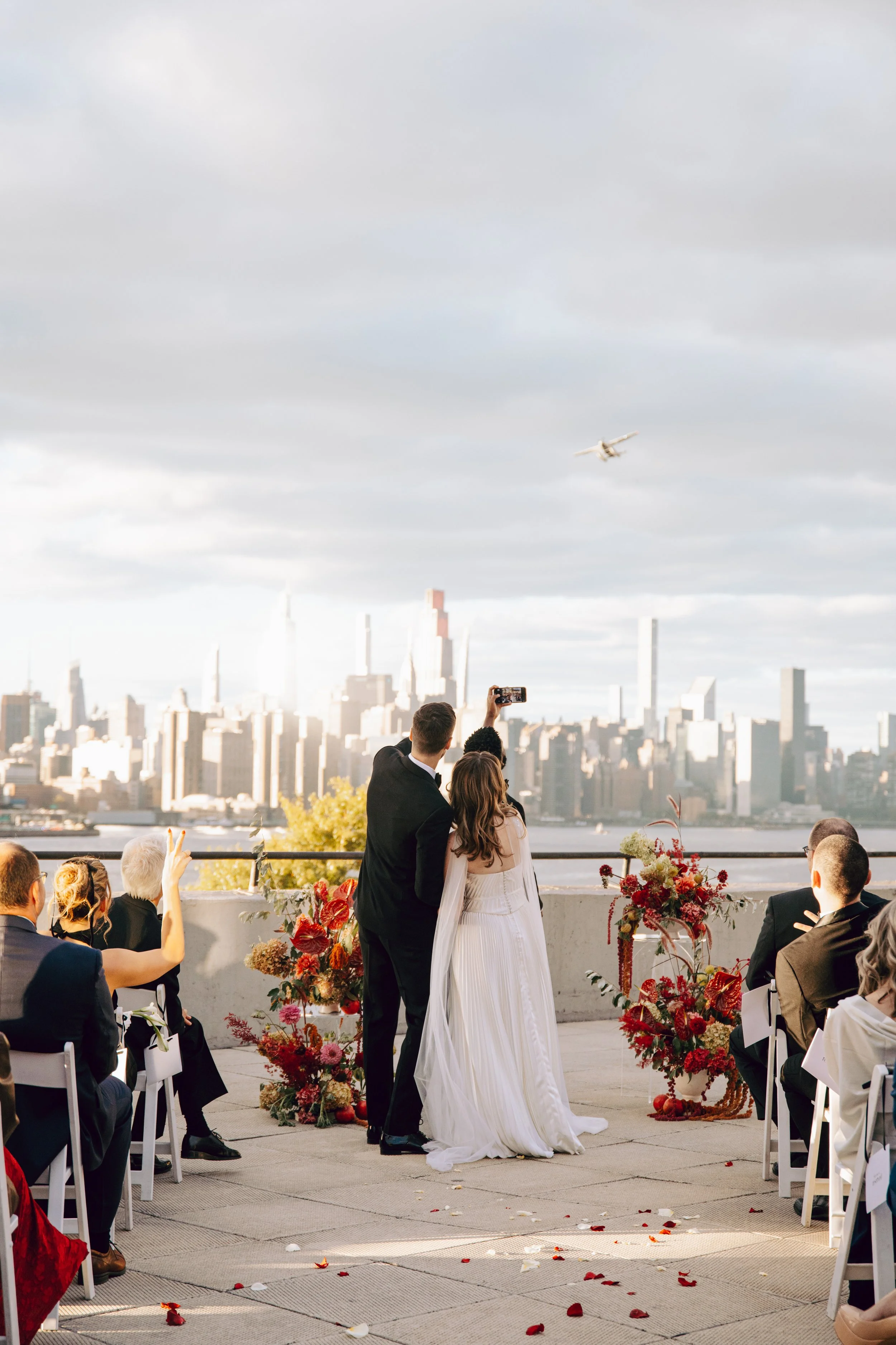 A bride and groom standing together at their wedding ceremony with a city skyline in the background. The bride is in a white gown, and the groom by her side, facing the city. Guests are seated and taking photos. Decorations with flowers and rose peta