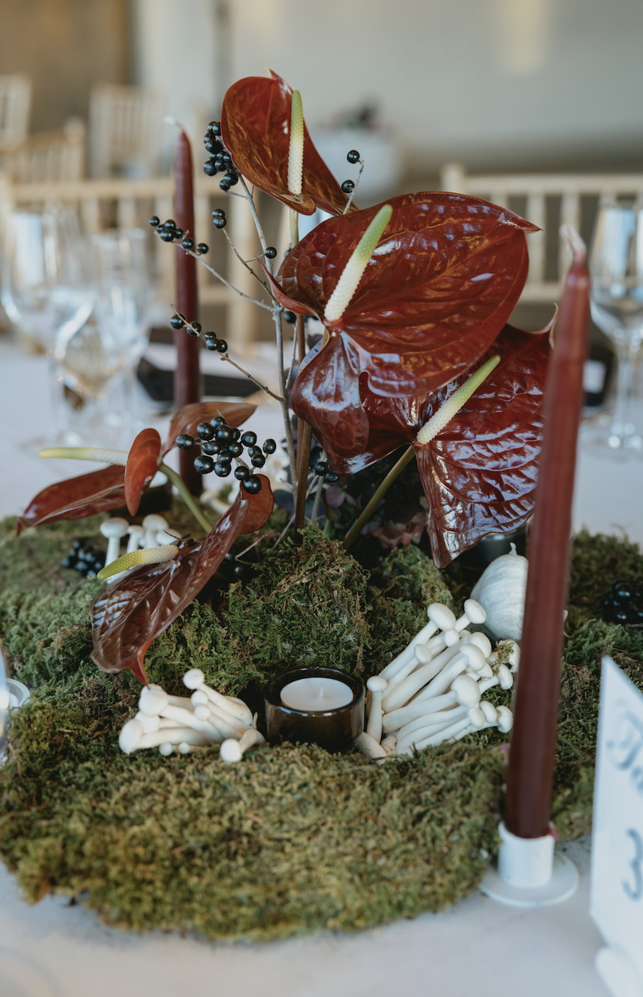 Decorative centerpiece with red anthurium flowers, black berries, white mushrooms, moss, and candles on a dining table.  marigolds florals nyc florist wedding florals the garrison
