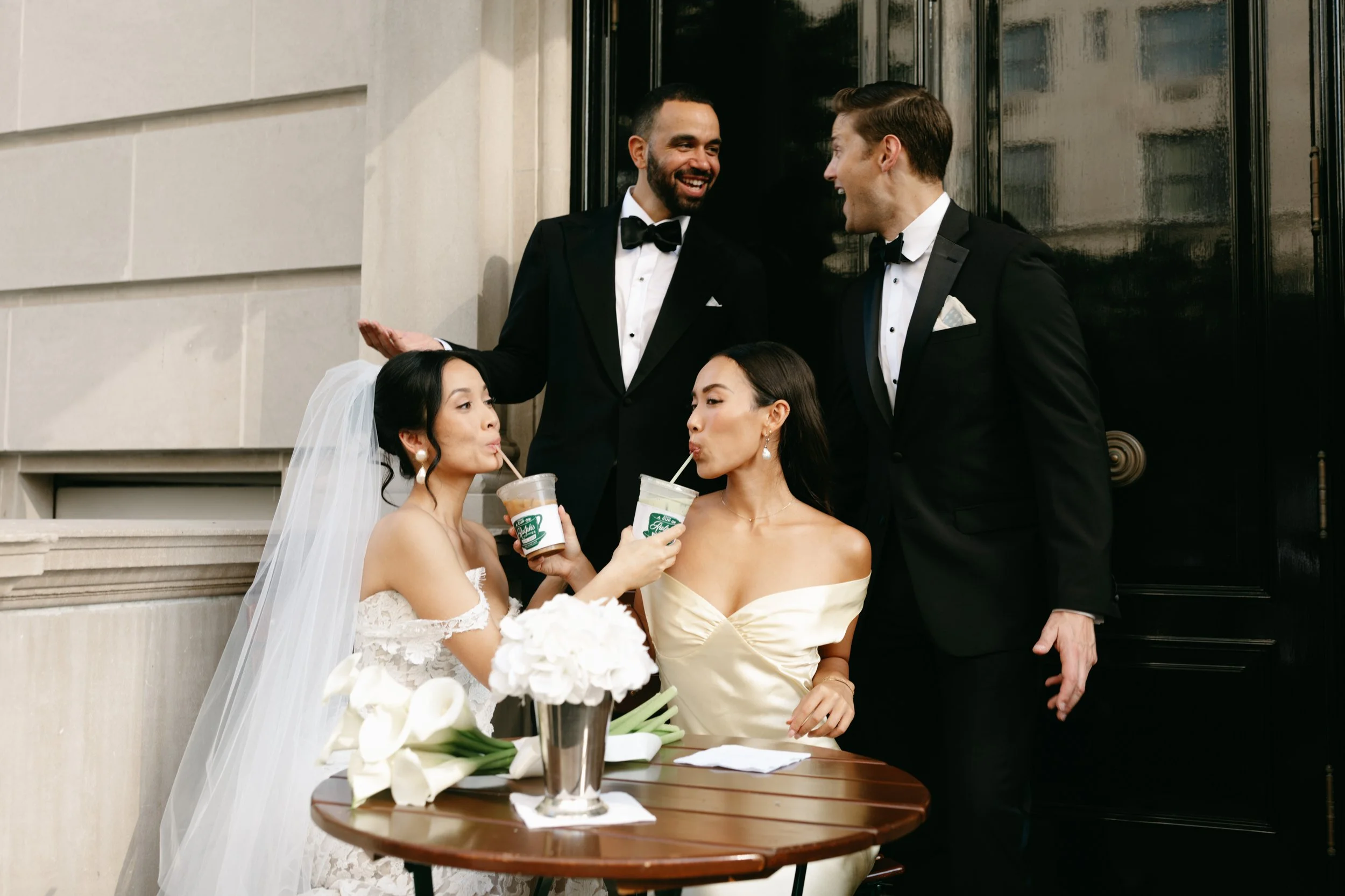 A group of two men and two women dressed in wedding attire outside a building, enjoying drinks together. marigolds florals nyc florist wedding florals central park boathouse wedding