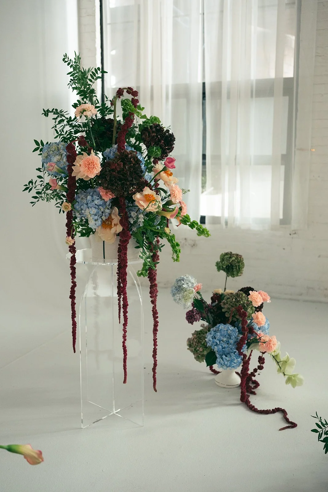 Two floral arrangements featuring hydrangeas, roses, and greenery, placed on a white table in front of a window with sheer curtains. marigolds florals nyc florist wedding florals atmosfera wedding