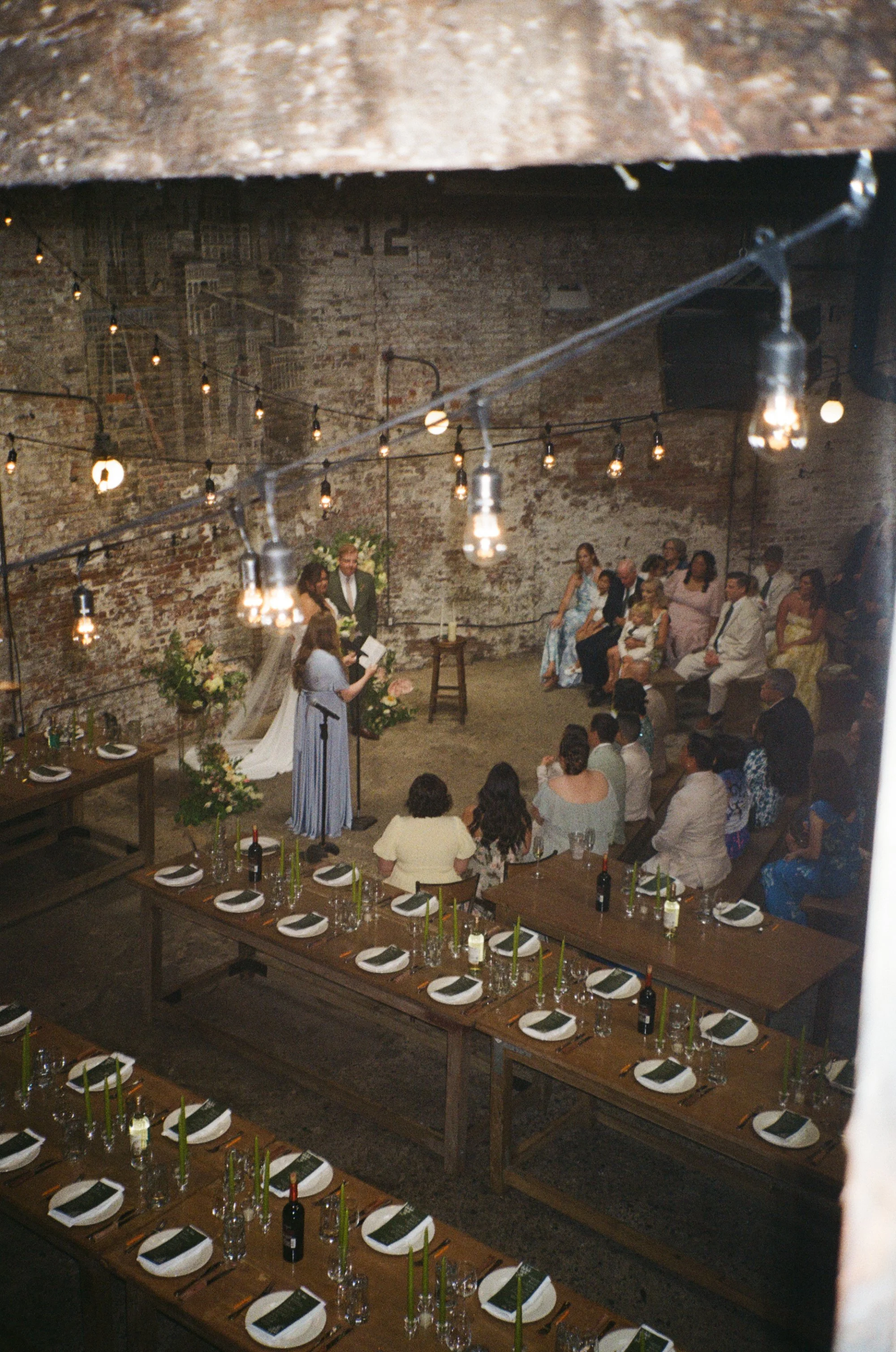 An indoor wedding reception setup with long wooden tables arranged with white plates, black napkins, wine bottles, and candles, in a rustic brick-walled venue. Guests are seated and a bride and groom are standing at the front during a ceremony, with 