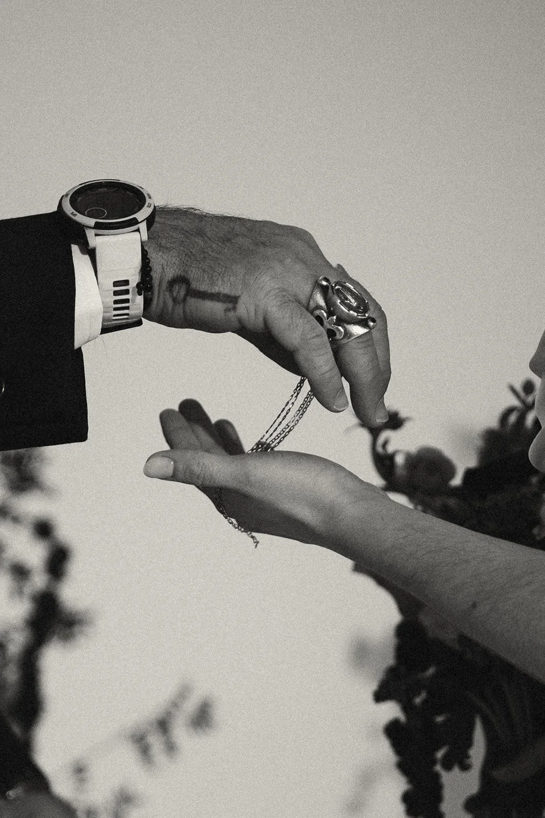 A black and white close-up of a handshake between two people, one wearing a watch and rings, the other with delicate jewelry, with blurred floral elements in the foreground. marigolds florals nyc florist wedding florals atmosfera wedding