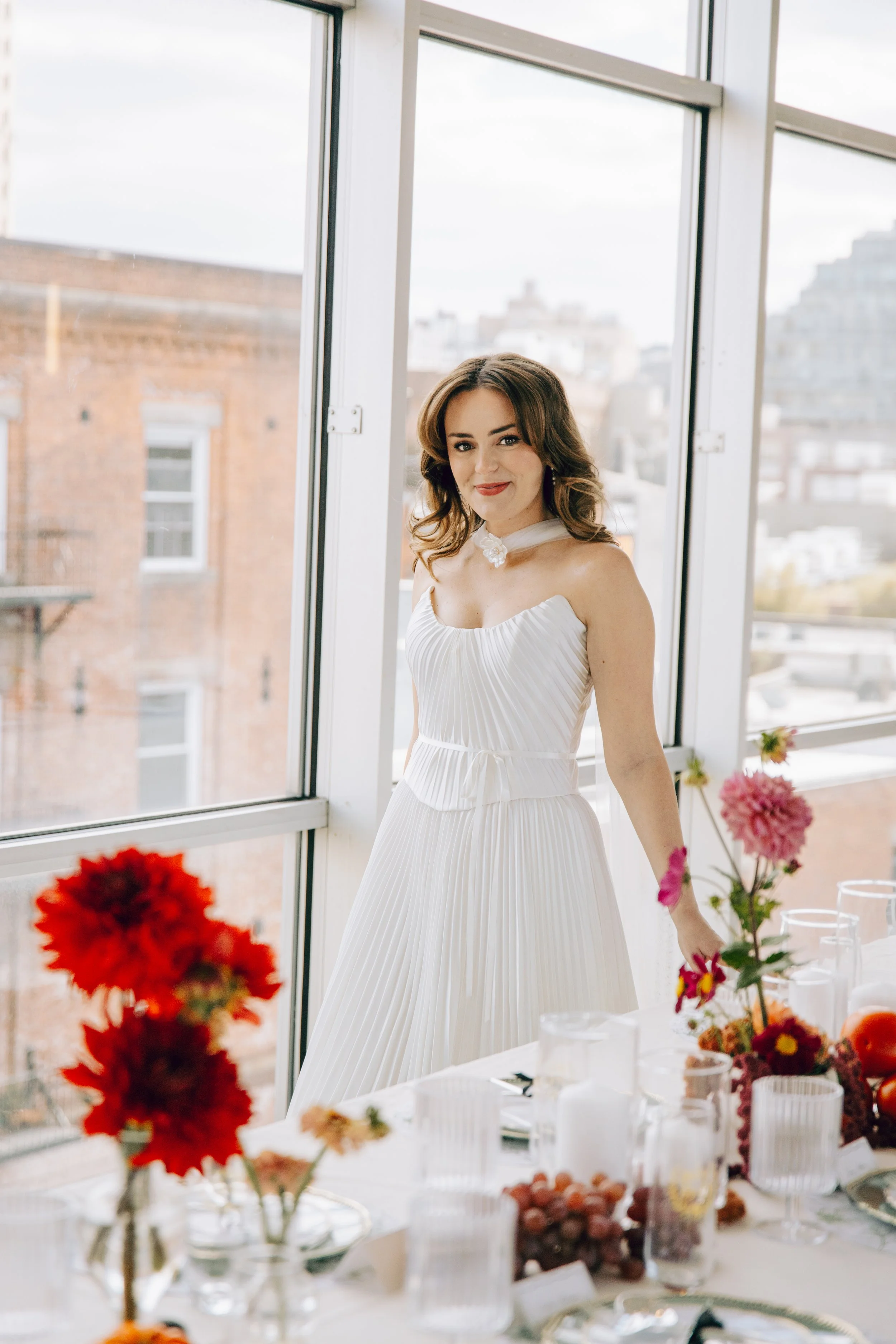 A young woman in a white pleated dress sits at a banquet table near a large window, with floral arrangements and tableware in the foreground, during daytime. marigolds florals nyc florist wedding florals the w loft wedding pedestal wedding ceremony 