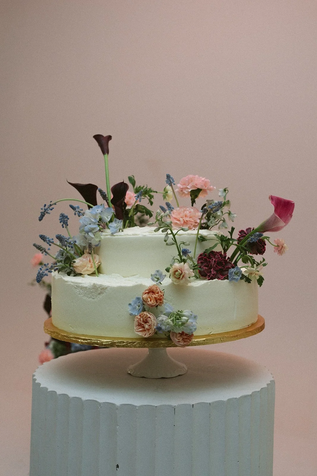 A white wedding cake decorated with a variety of pink, purple, and blue flowers, on a gold cake board, displayed on a white pedestal against a light pink background. marigolds florals nyc florist wedding florals atmosfera wedding
