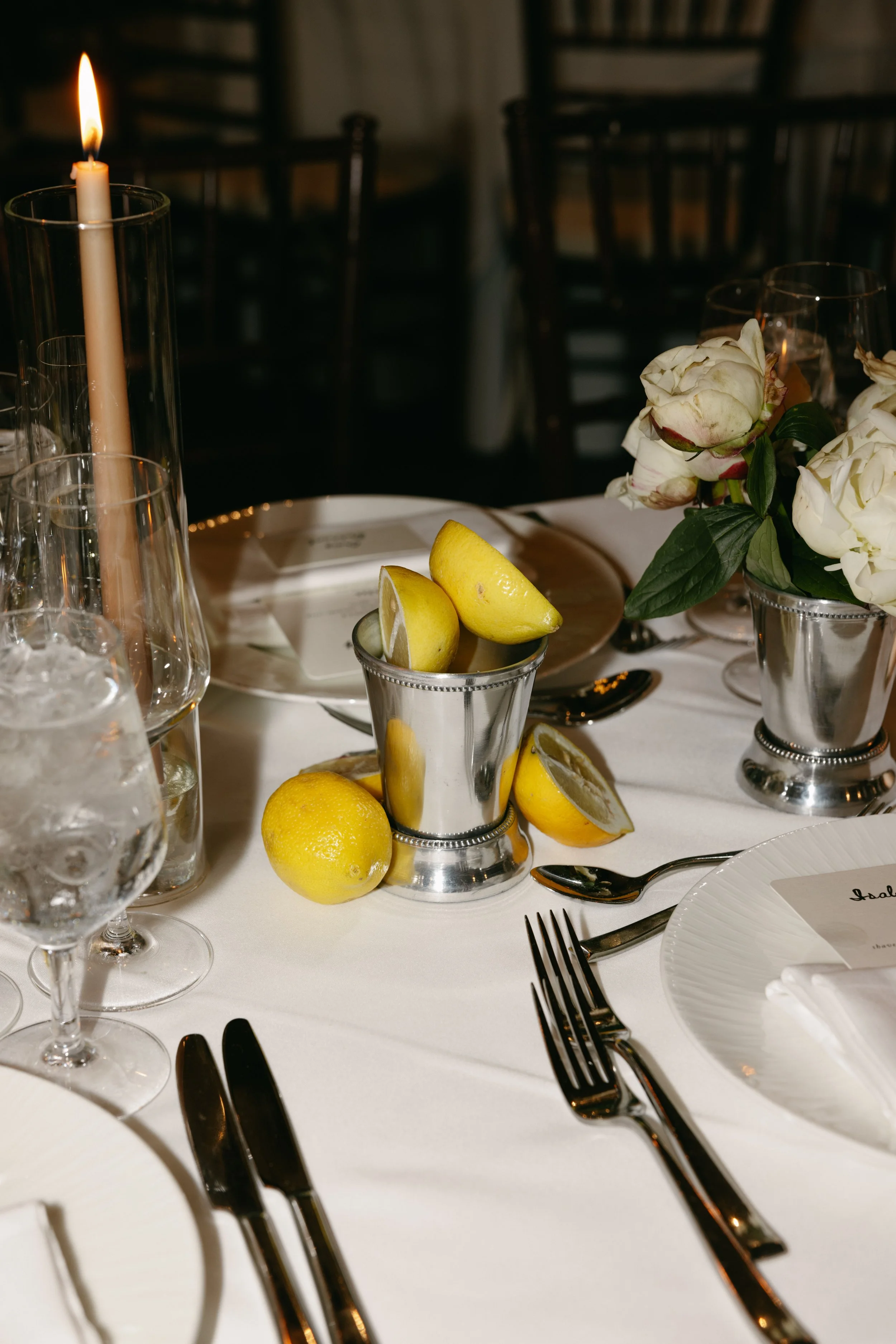 A formal dining table set with white tablecloth, silverware, wine glasses, a small bouquet of white flowers in a silver vase, a cluster of lemons, some cut lemon slices, tall candles, and a menu card.