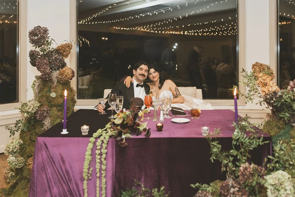 A couple dressed as a groom in a tuxedo and a bride in a wedding gown sitting at a decorated table with candles, glasses, and flowers, in front of a large window with string lights reflections. marigolds florals nyc florist wedding florals
