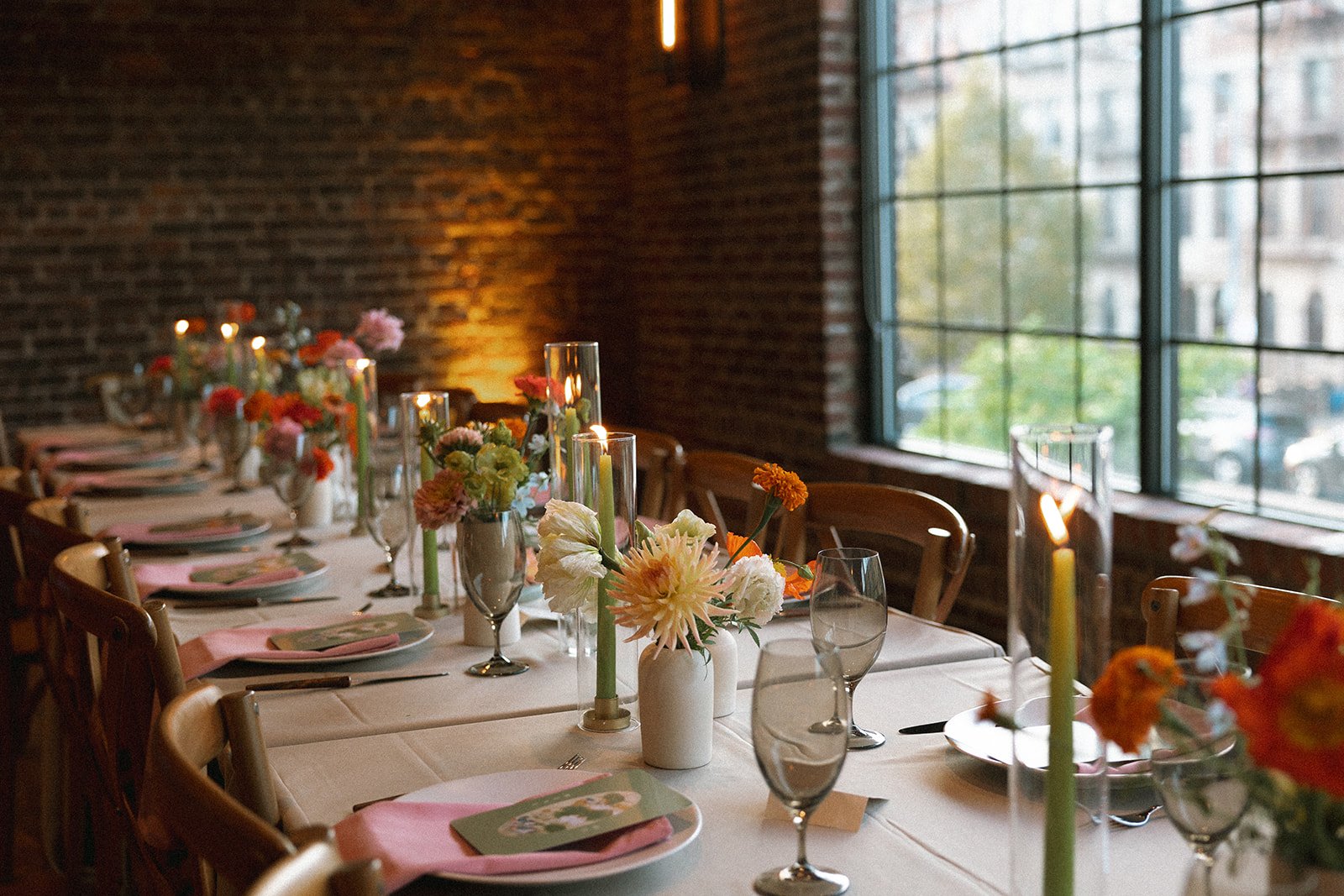 A long dining table decorated with floral centerpieces, candles, and place settings inside a room with brick walls and large windows. marigolds florals nyc florist wedding florals