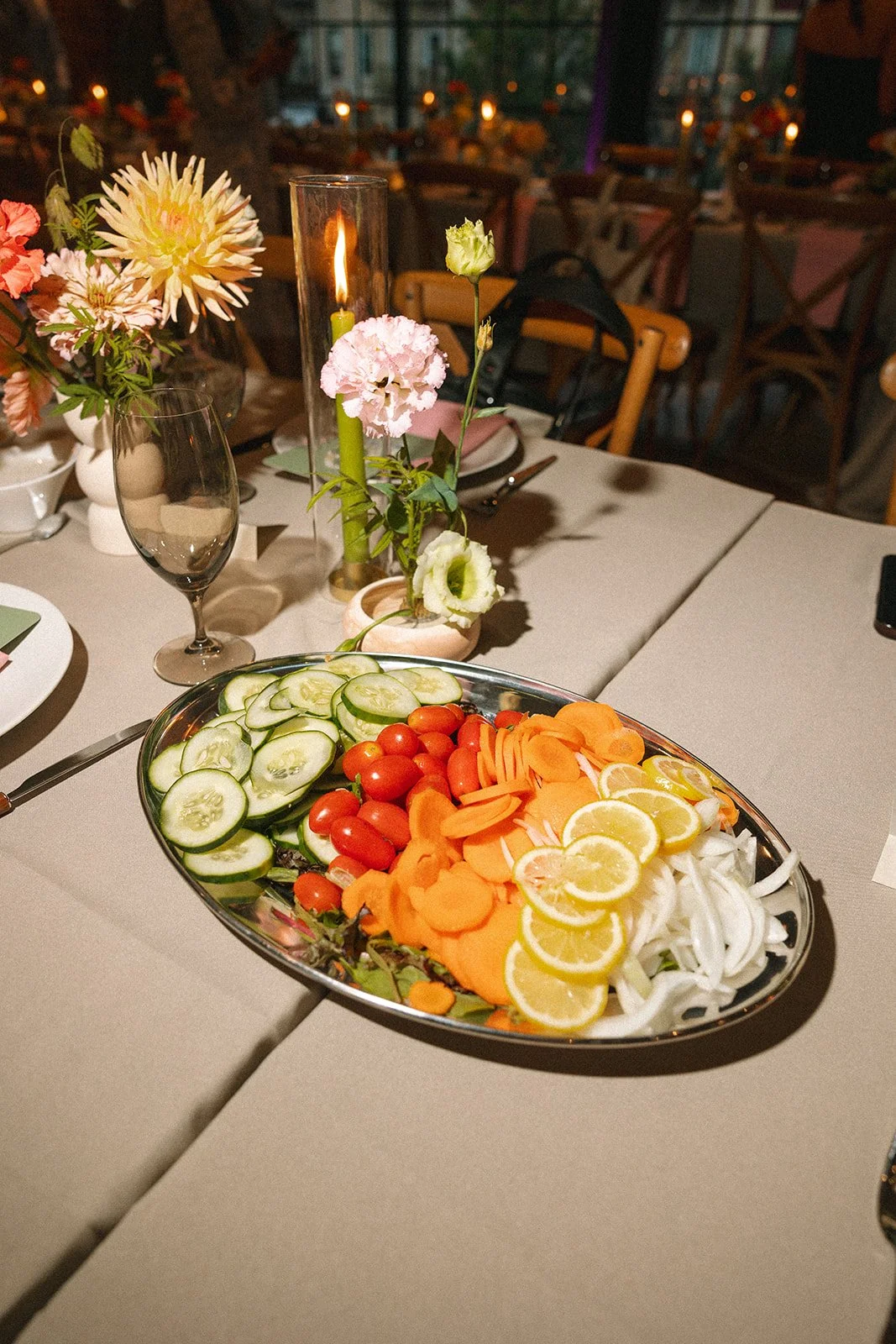 A table set for dinner with a centerpiece of flowers and a lit candle, along with a platter of sliced cucumbers, cherry tomatoes, sliced carrots, and lemon wedges, in a cozy restaurant setting. marigolds florals nyc florist wedding florals