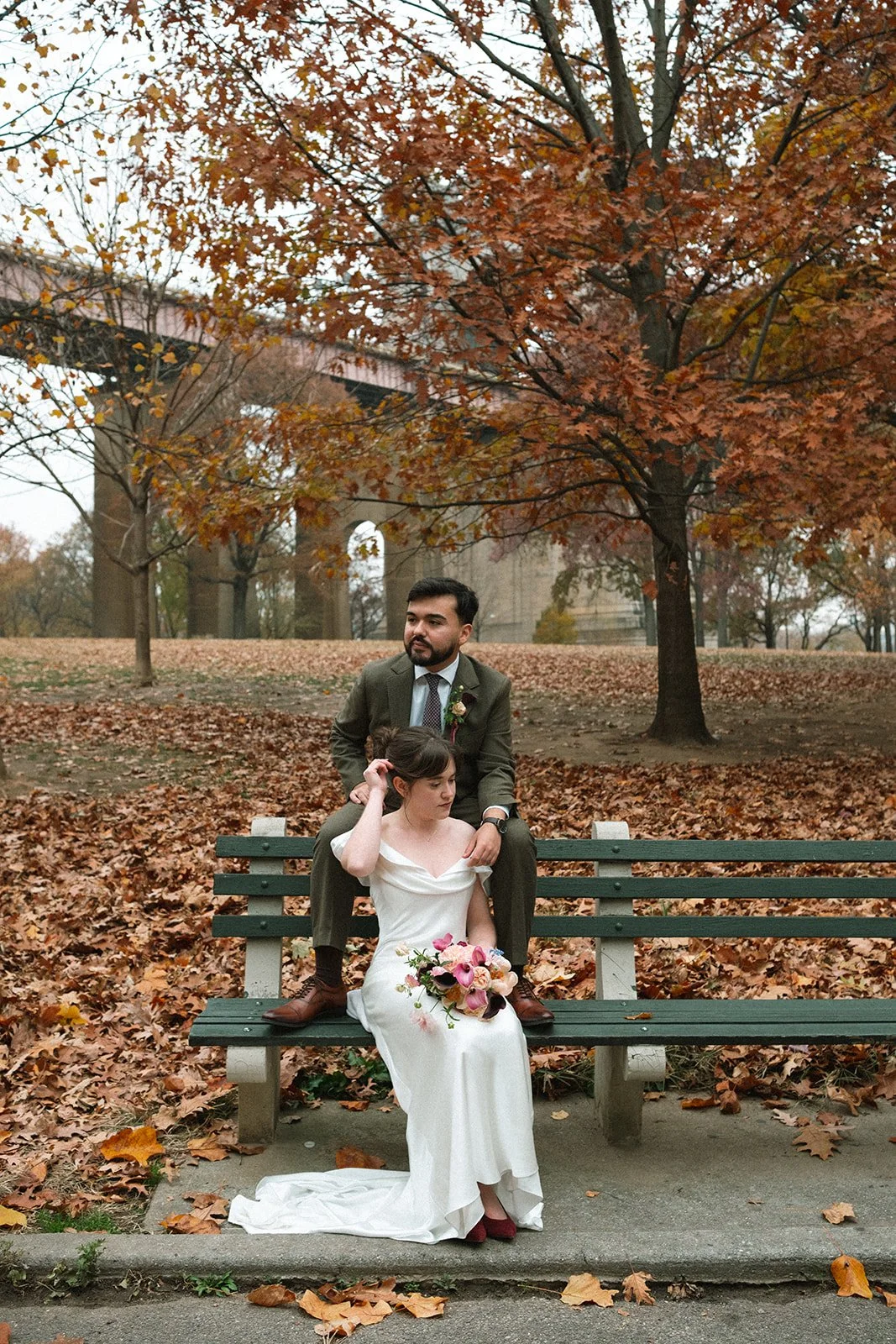 A bride and groom in wedding attire sitting on a park bench during autumn, surrounded by fall leaves and colorful trees. marigolds florals nyc florist wedding florals atmosfera wedding