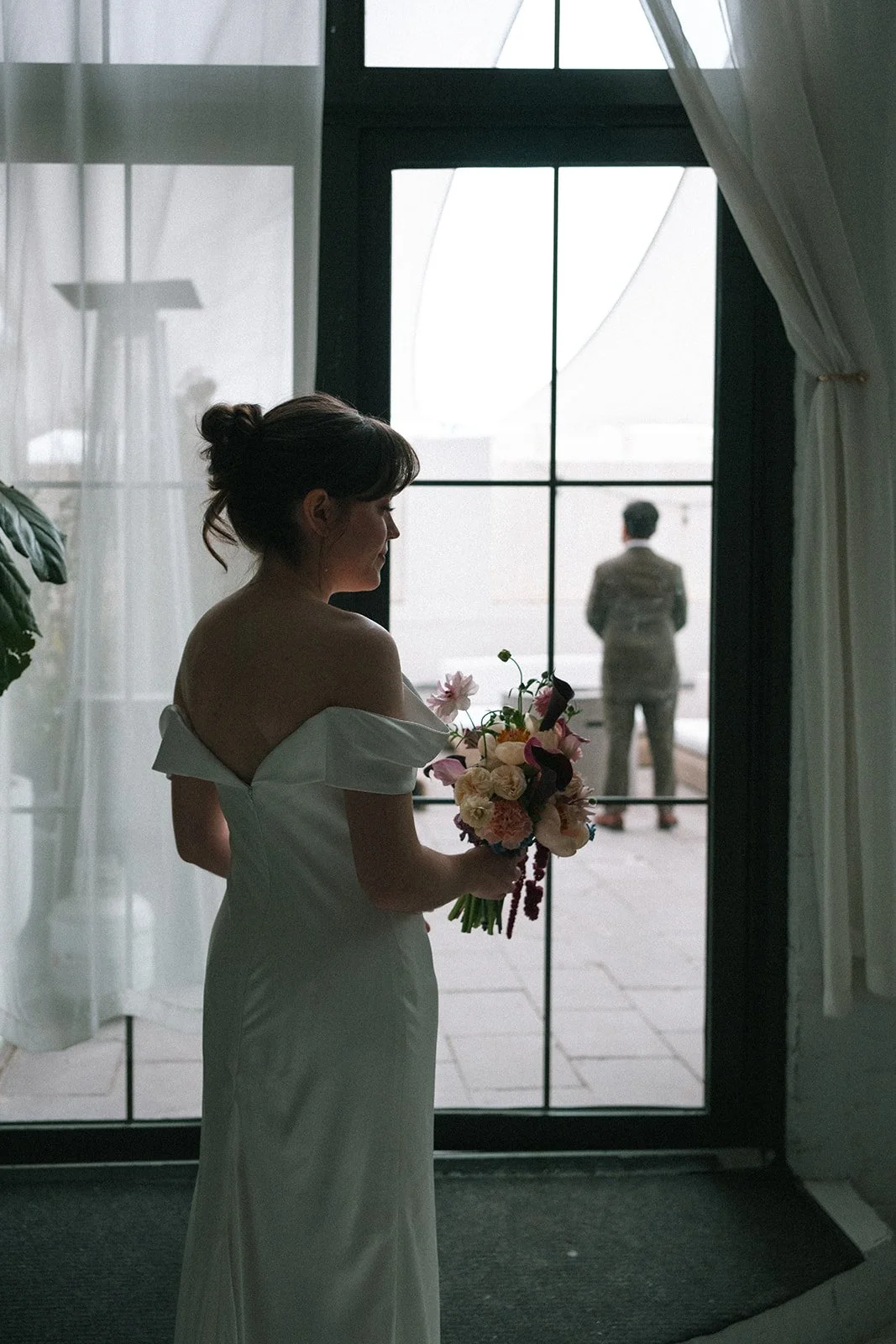 A woman in a white off-shoulder dress holding a bouquet of flowers, looking thoughtfully out a window. A man stands outside, facing away, in a suit. marigolds florals nyc florist wedding florals atmosfera wedding