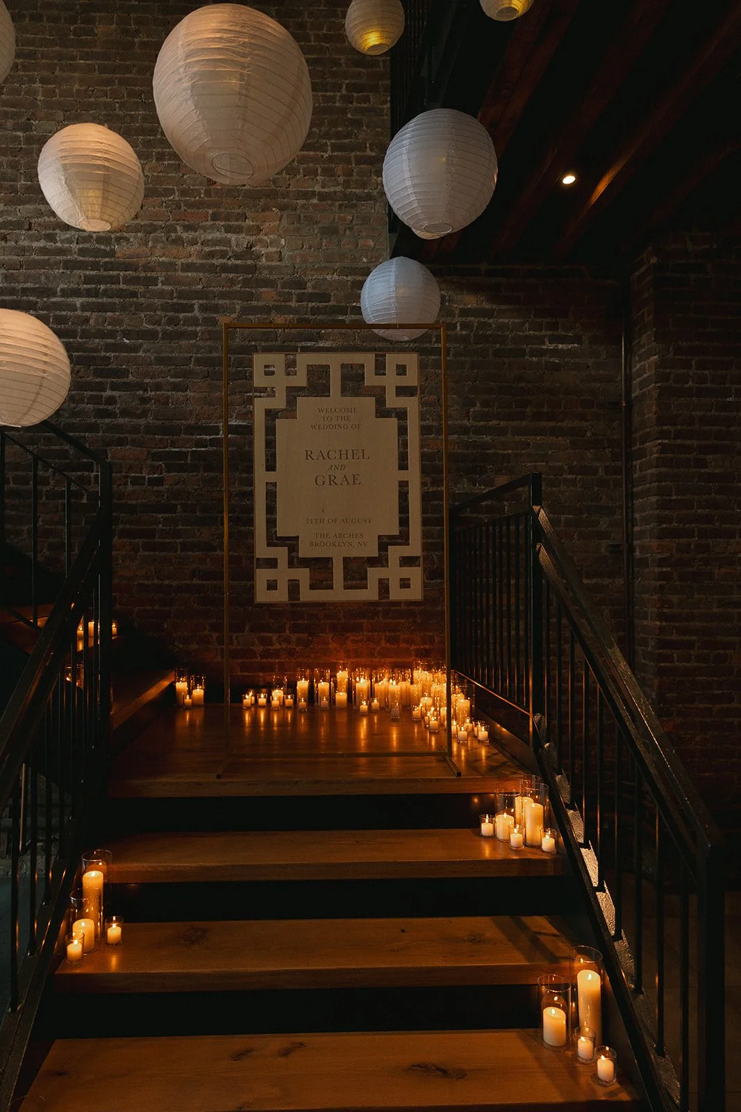 A staircase with black railing and wooden steps, decorated with numerous candles in glass holders. On the brick wall at the top of the stairs, there is a wedding welcome sign for Rachel and Grae, dated August 21st, at The Arches in Brooklyn, NY. Seve