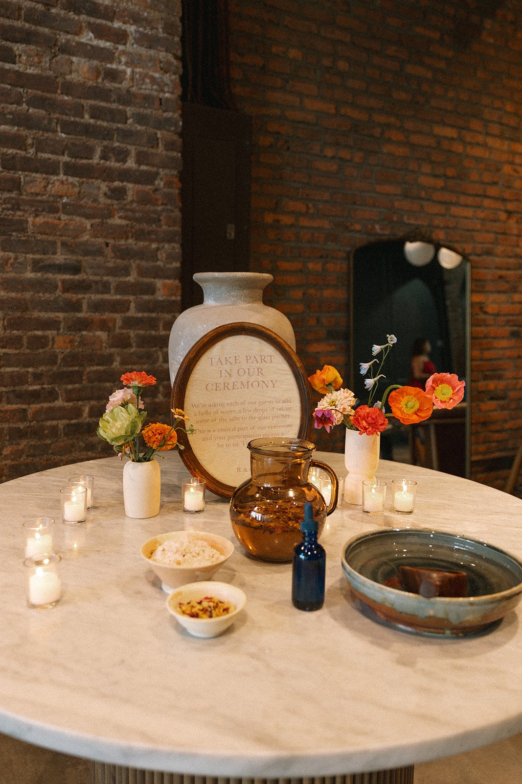 Round table decorated with flowers, candles, a framed sign, vases, a pitcher, bowls, and a dropper bottle, set against a brick wall in an indoor setting. marigolds florals