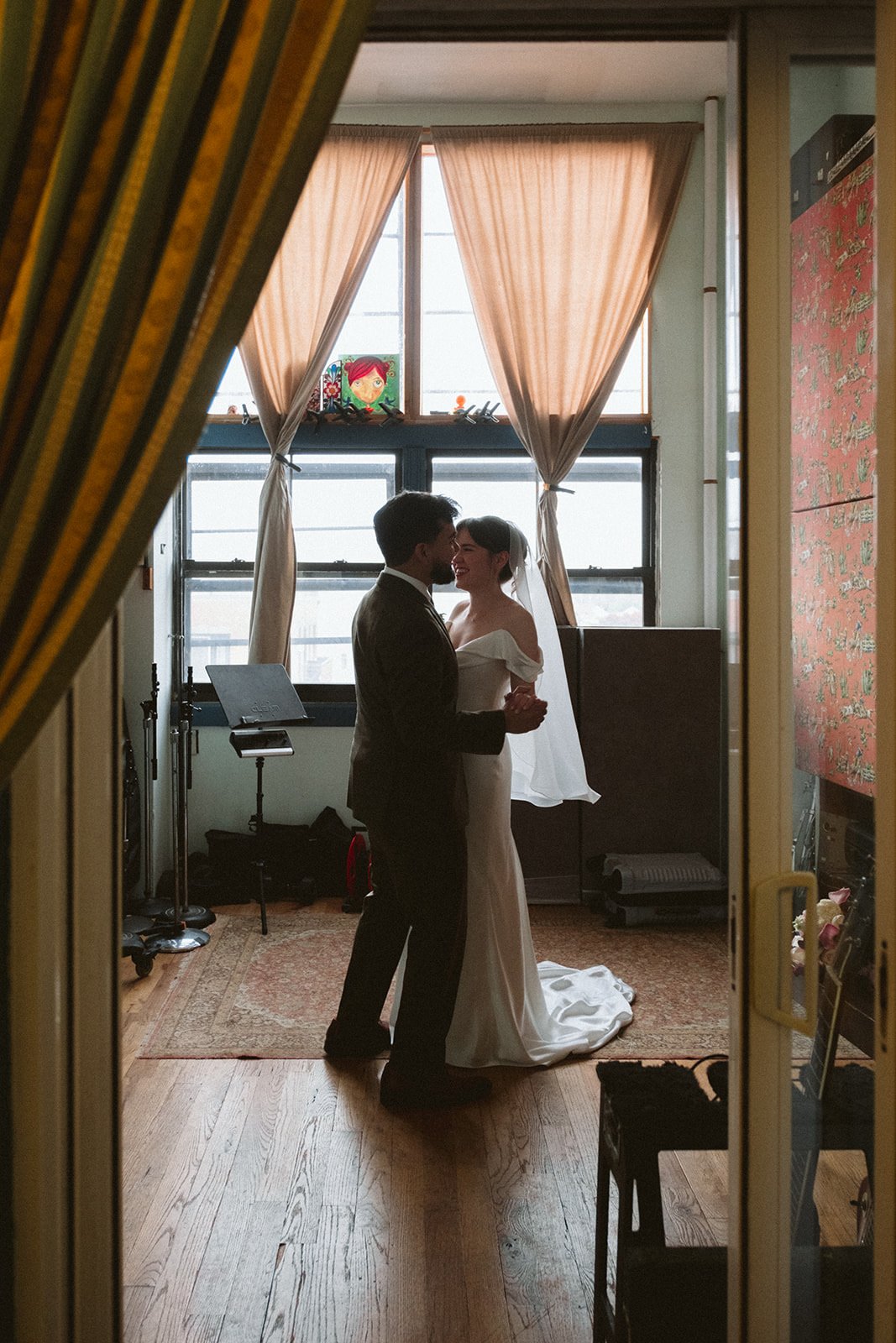 A bride and groom dancing inside a room with large window and beige curtains, seen through an open door with yellow framing. marigolds florals nyc florist wedding florals atmosfera wedding